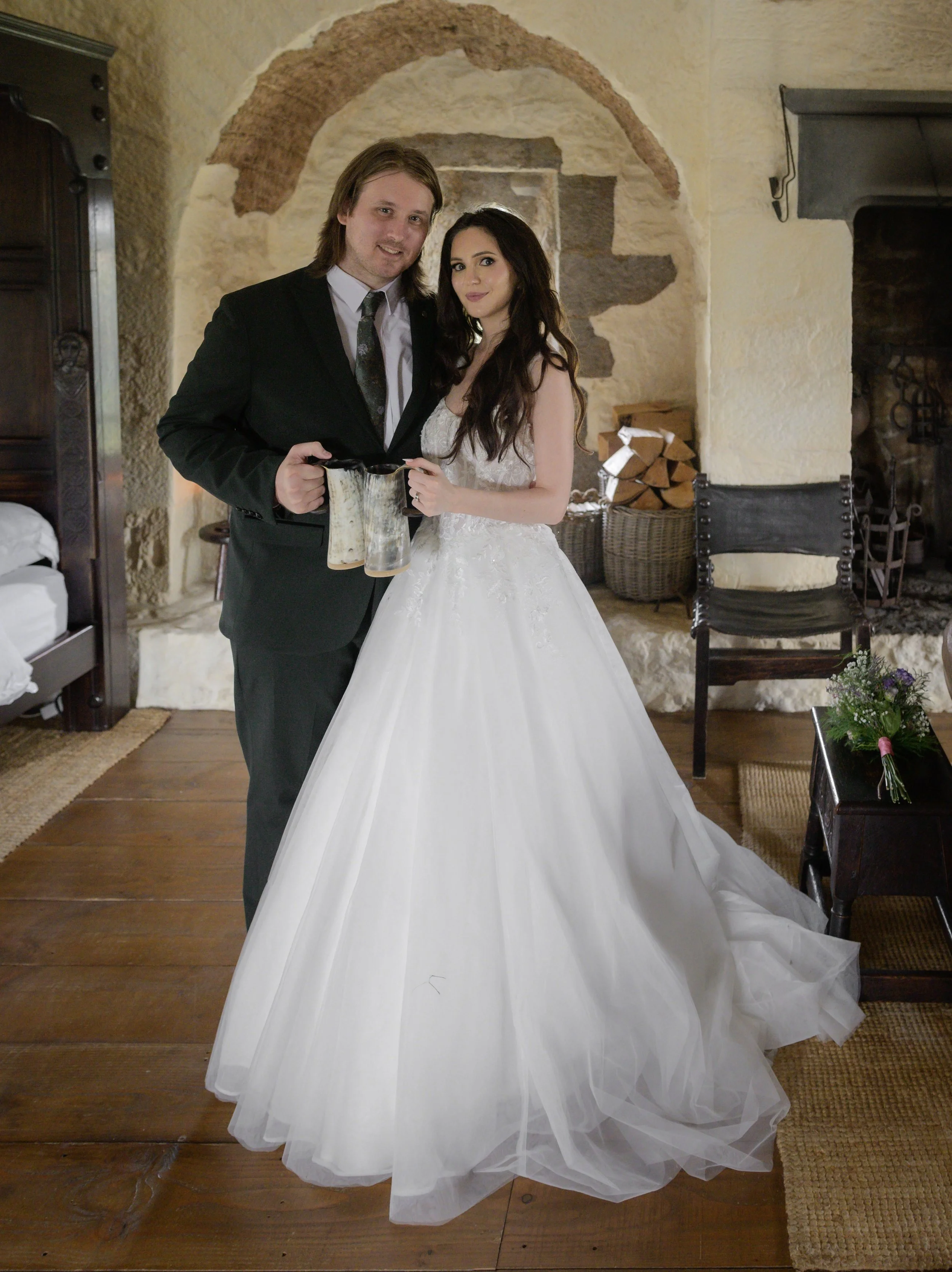 A bride and groom holding glasses in a cozy, rustic indoor setting with stone walls, wooden flooring, and a firewood basket.