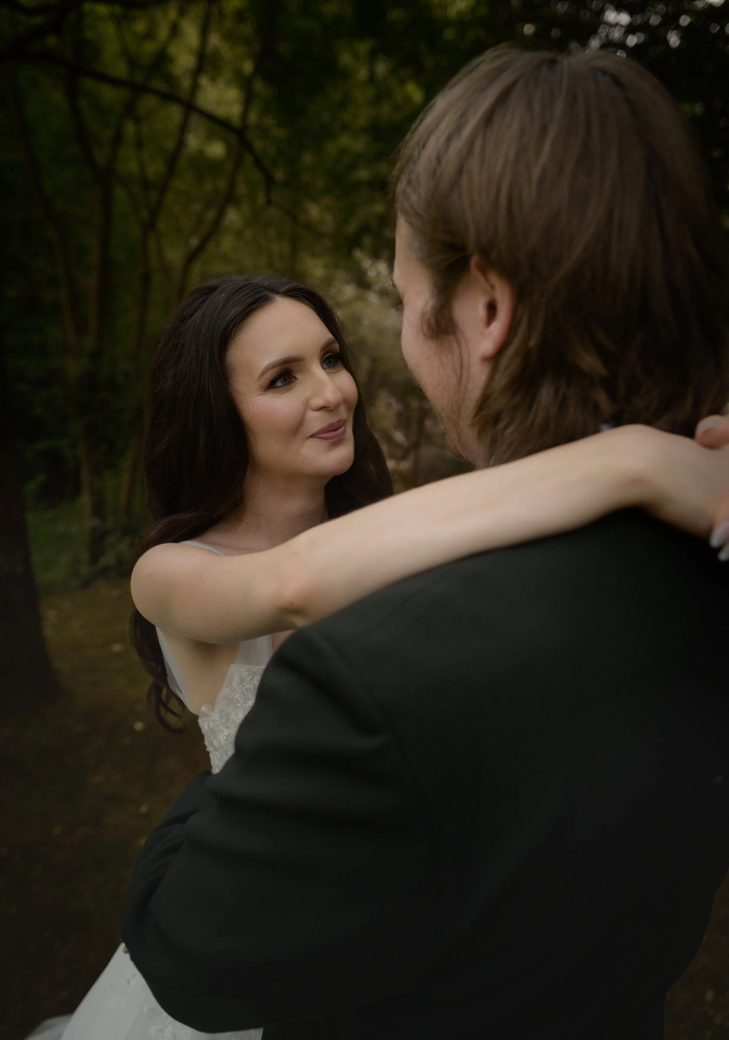A bride and groom are dancing outdoors in a wooded area, with the bride smiling and looking at the groom.