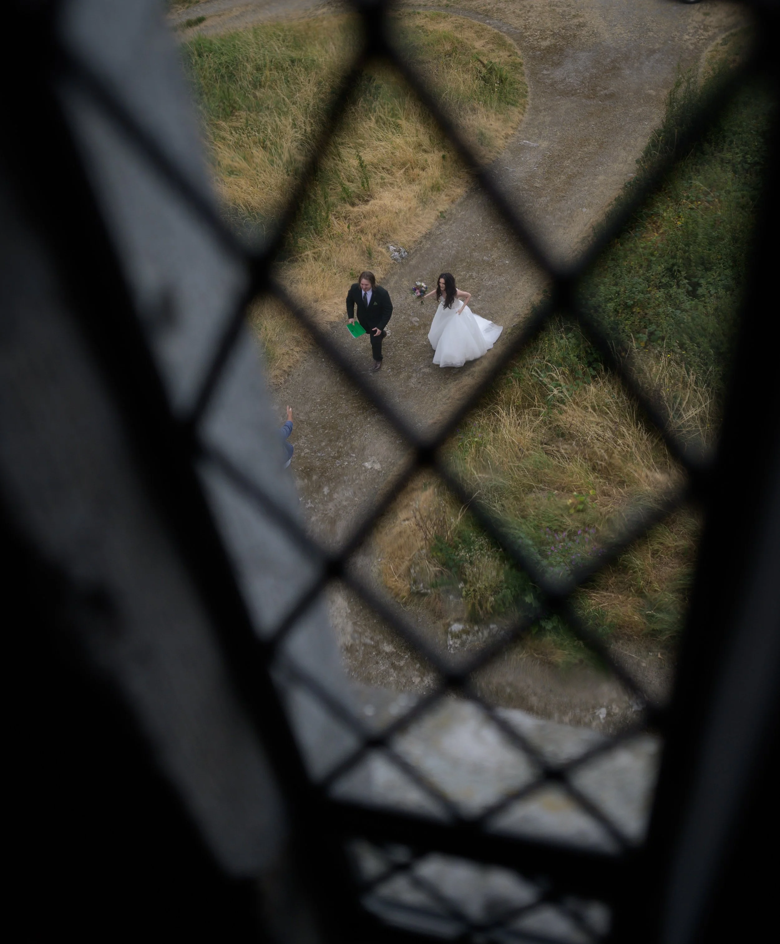 A couple dressed in wedding attire standing on a dirt path, viewed from above through a chain-link fence.