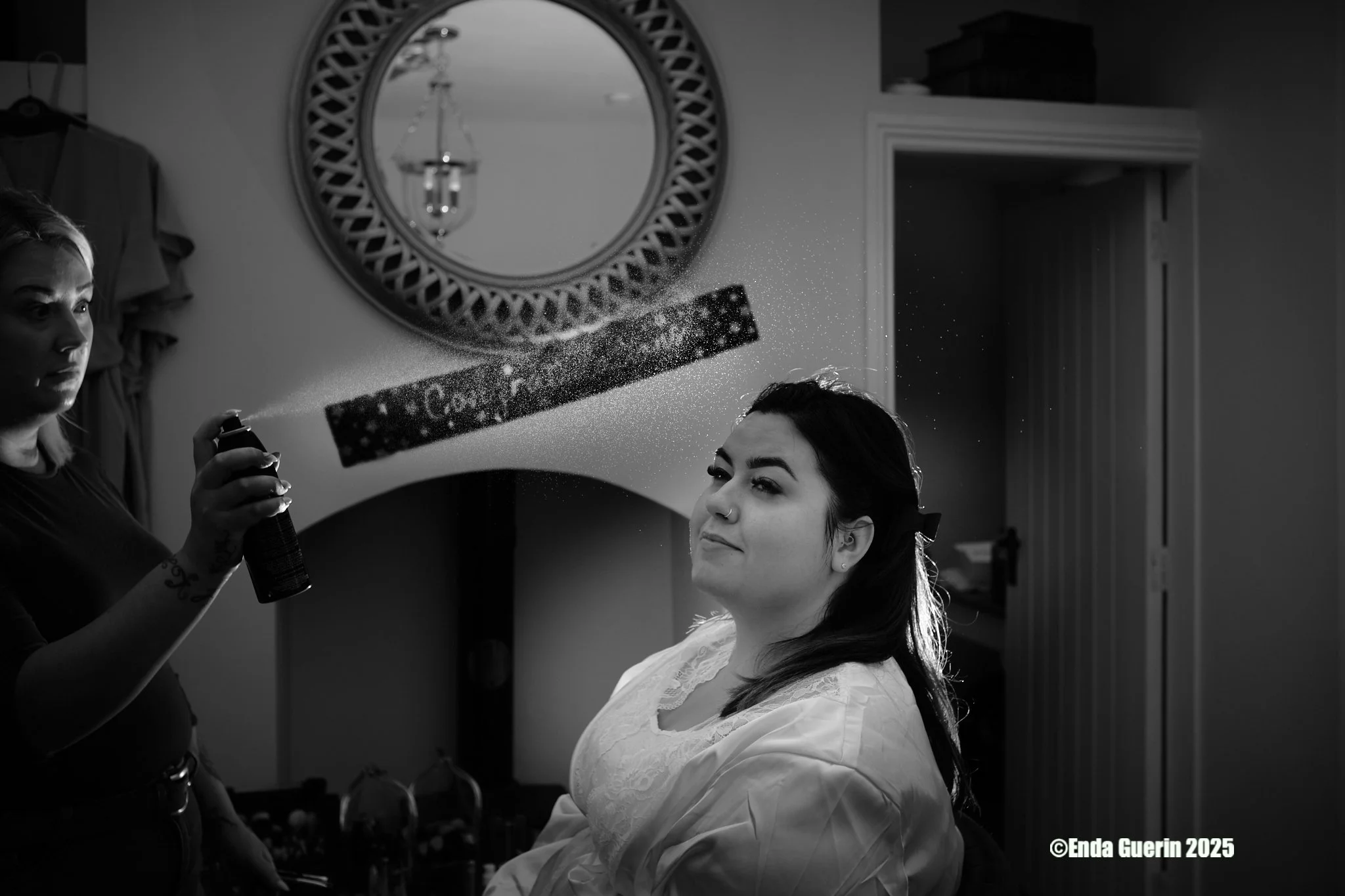 Woman sitting in a chair having her hair and makeup done, with a makeup spray being applied by a stylist. The stylist is on the left side holding a spray can. The woman is smiling slightly, wearing a white garment. A round mirror and a chandelier are