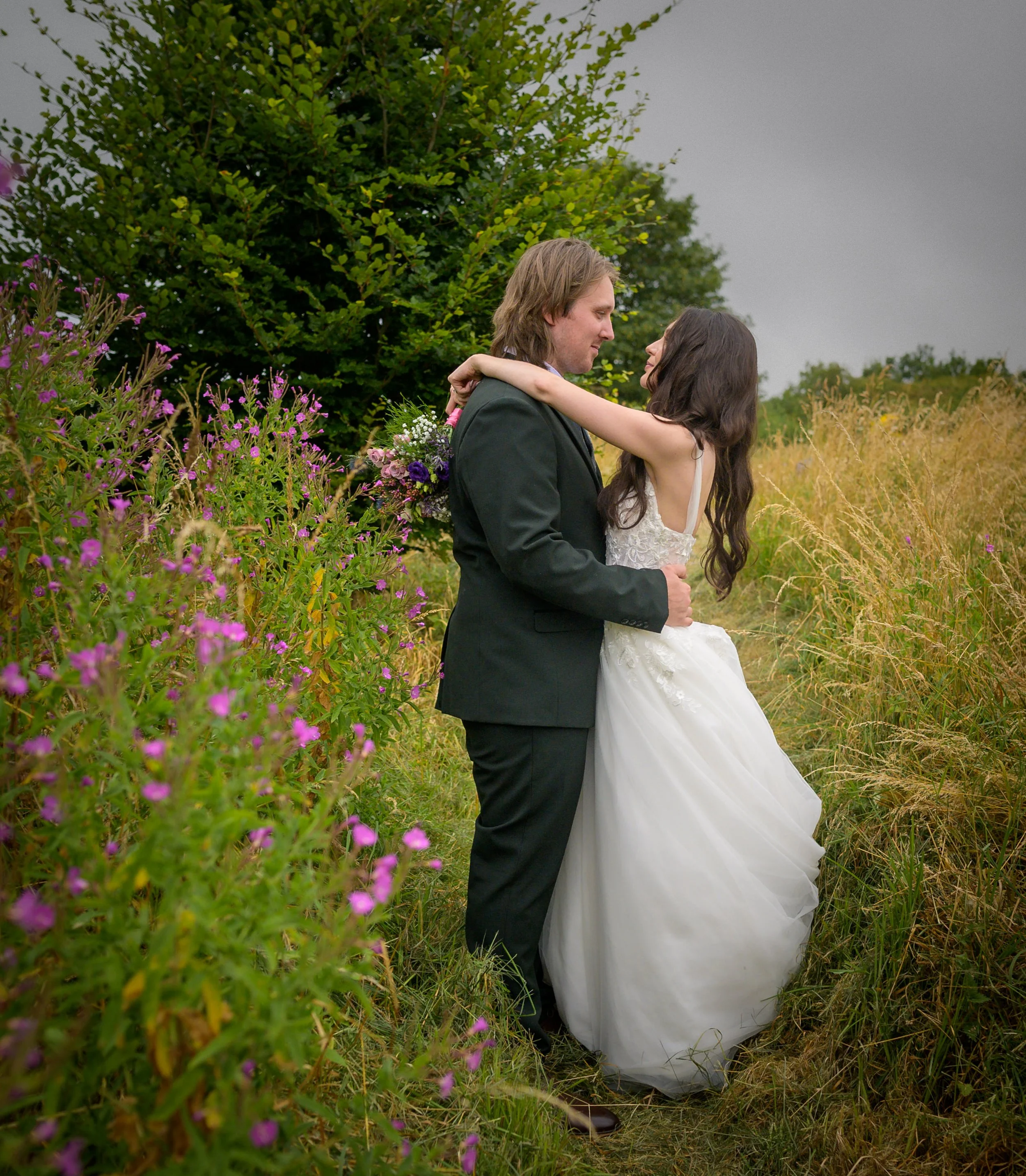 A couple embracing outdoors in a field with purple wildflowers and green trees in the background, with overcast sky.