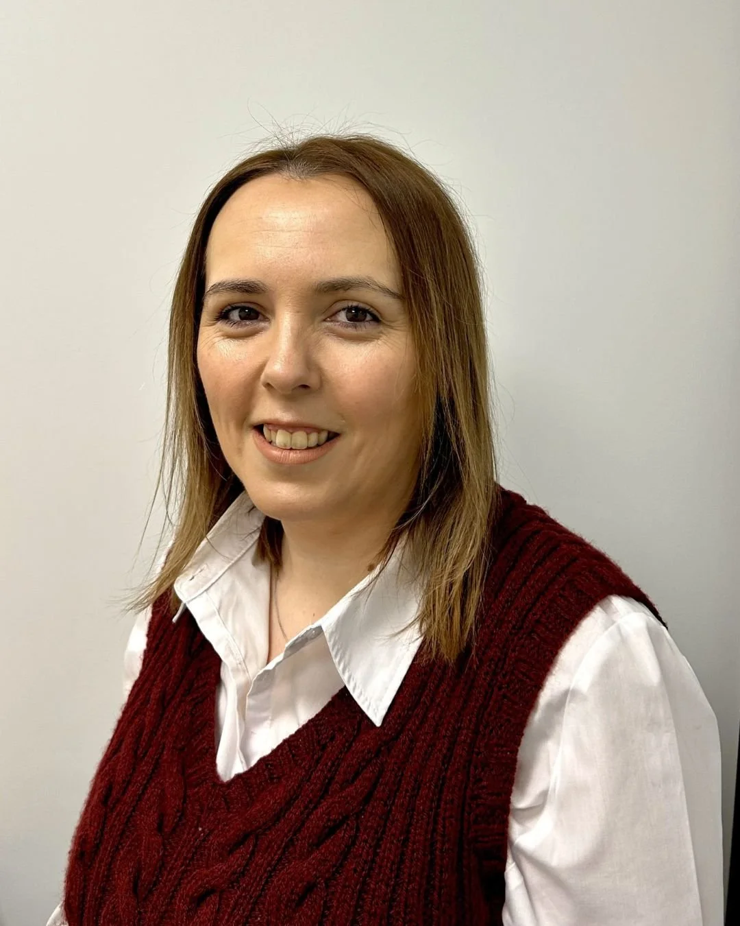 Our People & Culture Director at Coventry Building Society Arena, with shoulder-length brown hair, wearing a white shirt and a red knitted vest, smiling in front of a plain light-colored wall.