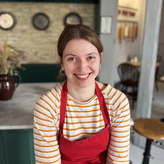 A young woman with a bright smile, wearing a red apron over a yellow and white striped shirt, sitting in a cafe or restaurant with a rustic background.