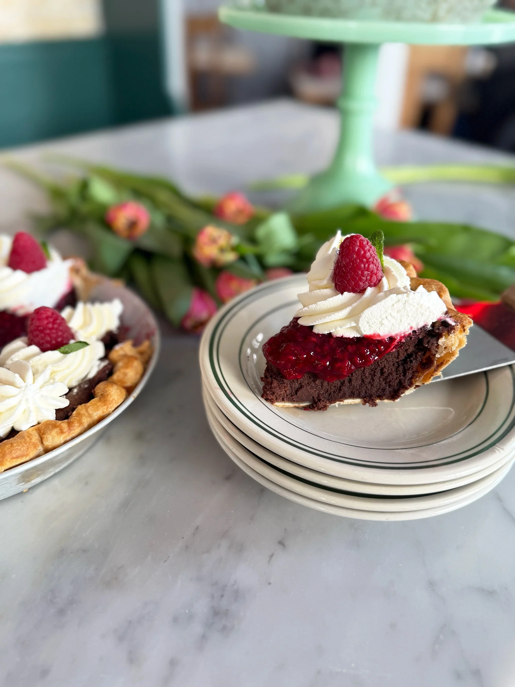 Close-up of a slice of chocolate berry pie topped with whipped cream and a raspberry, placed on a stack of white plates, with a blurred background of a table and a green cake stand.