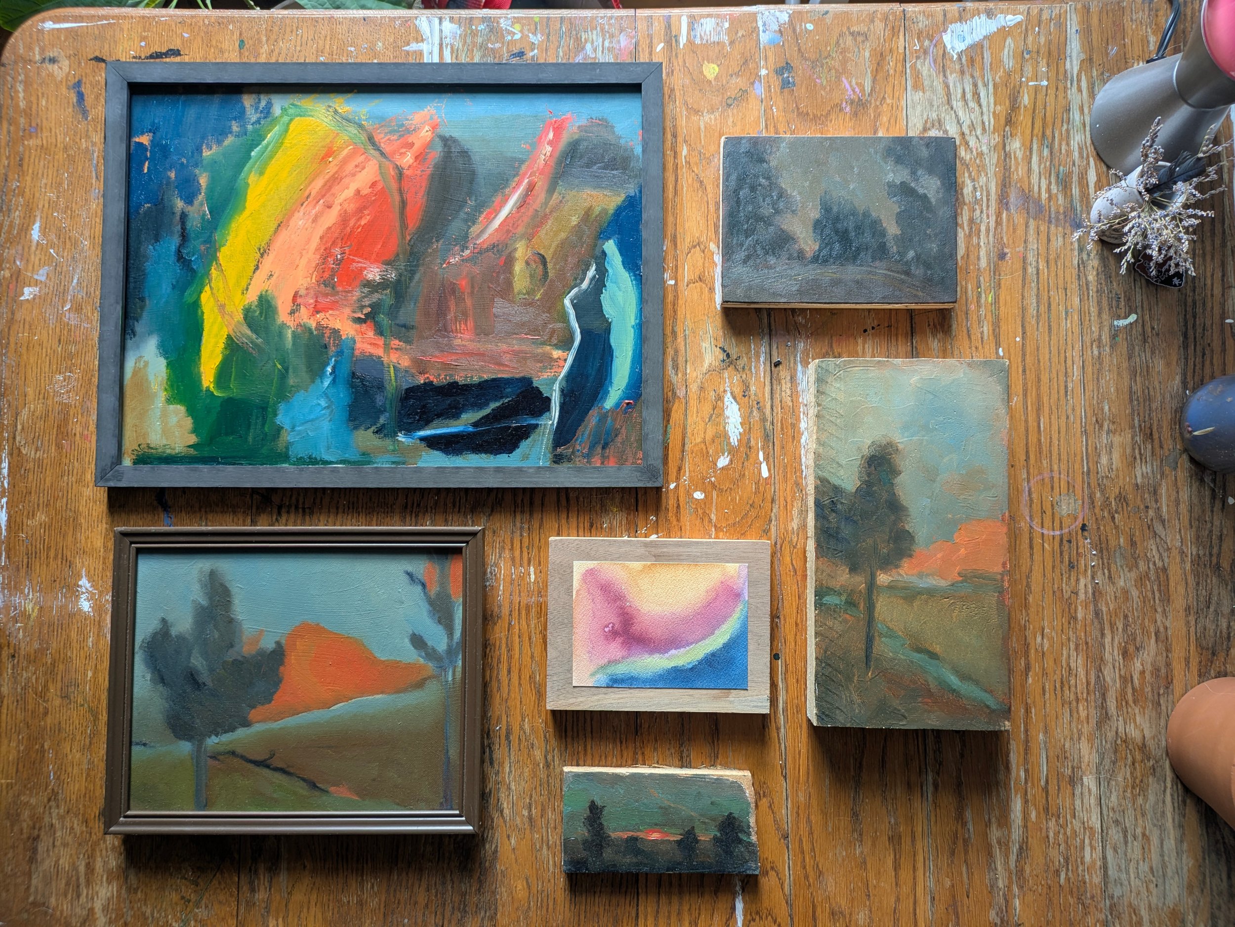 A wooden table with several small and large abstract paintings, a painted wood frame, and a metal water bottle with dried flowers.