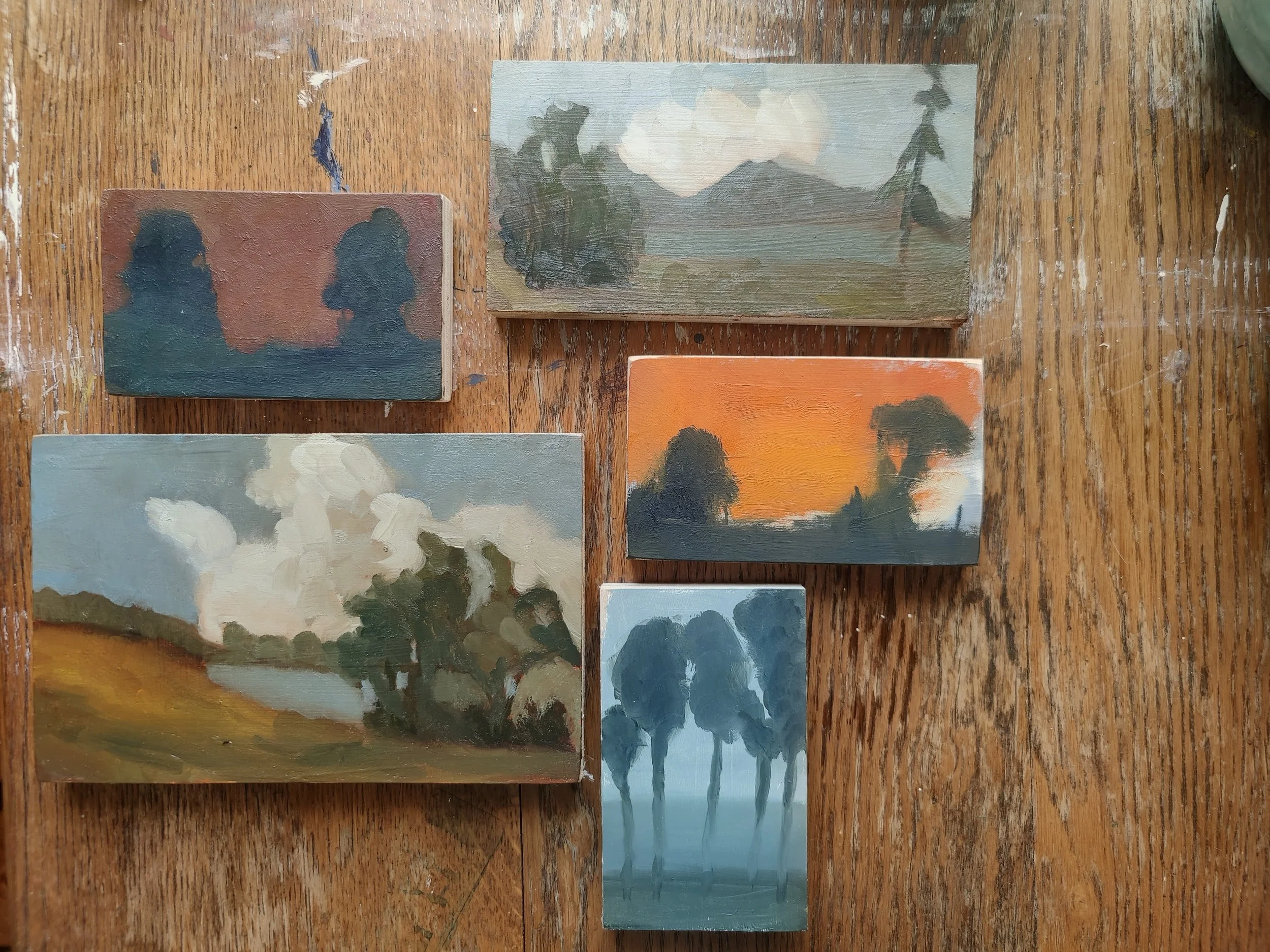 Six small landscape paintings placed on a wooden surface, depicting various nature scenes such as mountains, trees, and skies at different times of day.