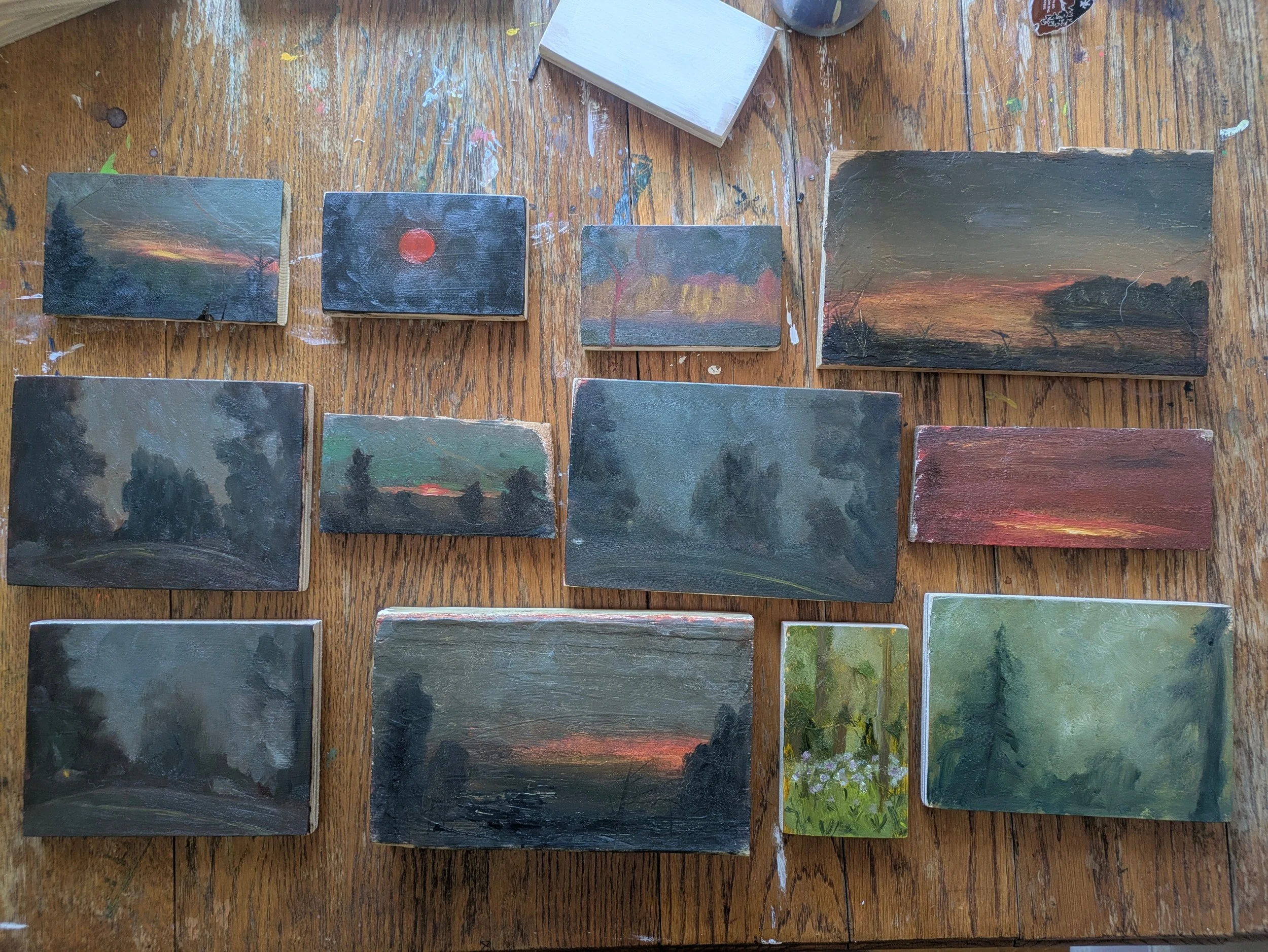 A collection of small landscape paintings on a wooden table, depicting various sunset and nature scenes, with some featuring trees and flowers.