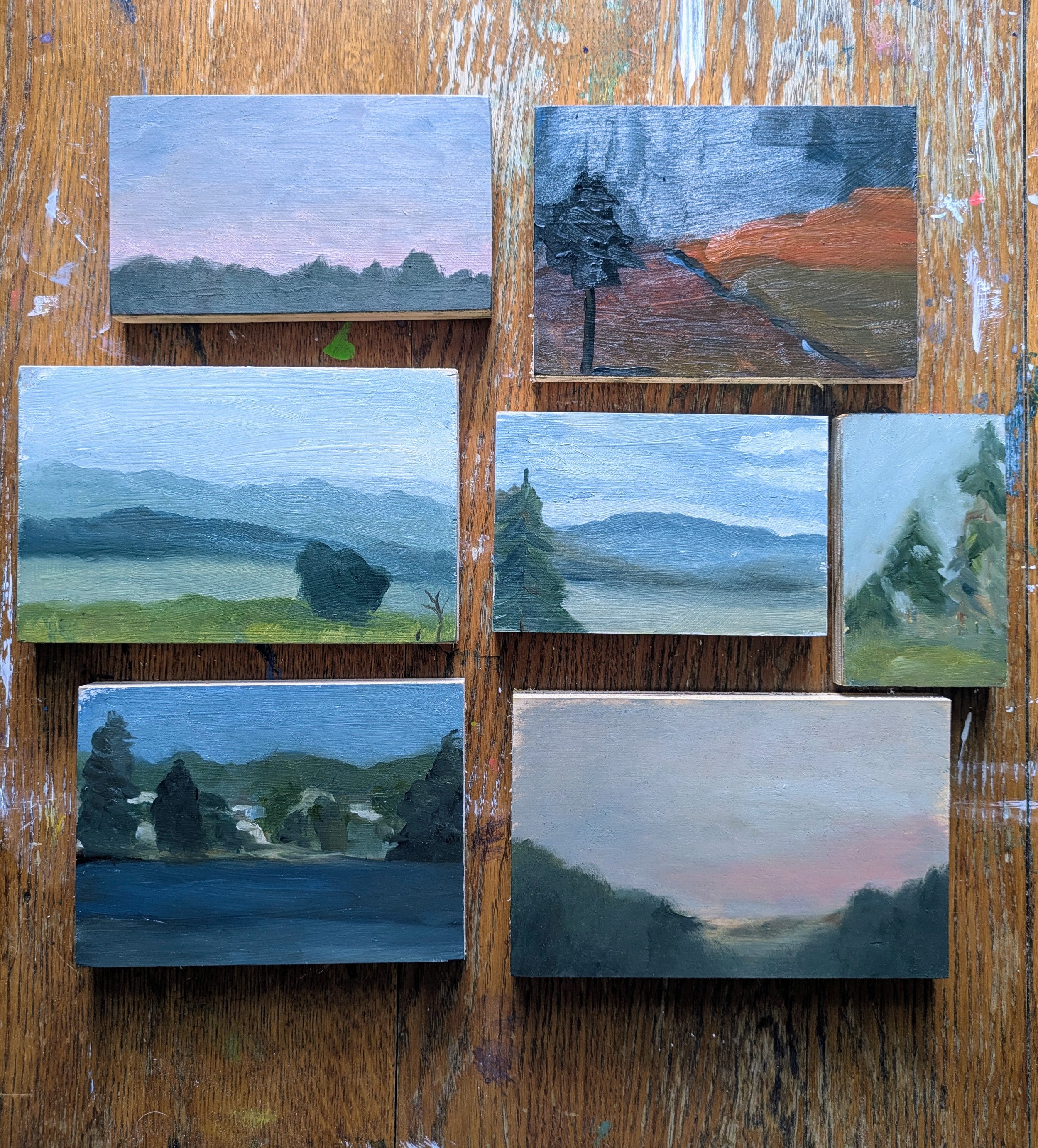 A set of five miniature landscape paintings on a wooden table, depicting various natural scenery such as mountains, mountains with trees, hills, and forests, painted with acrylic or oil paints.