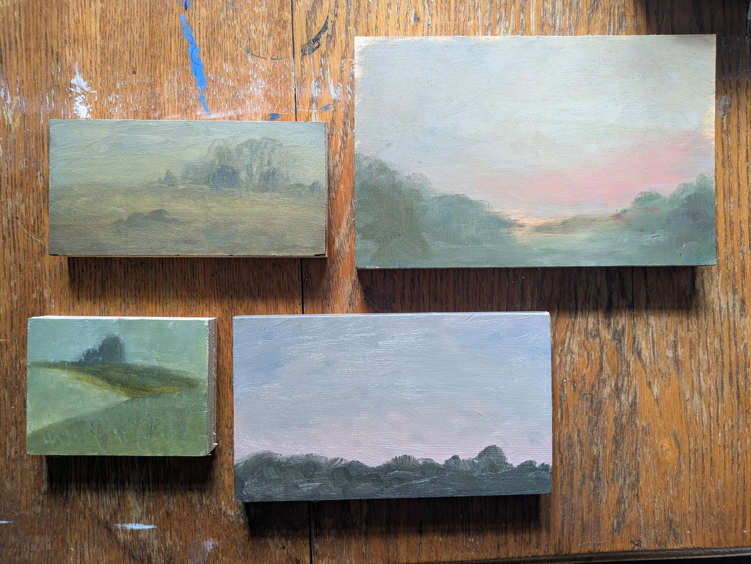 Multiple small landscape paintings on a wooden table, depicting misty countryside scenes with trees and sky in soft colors.
