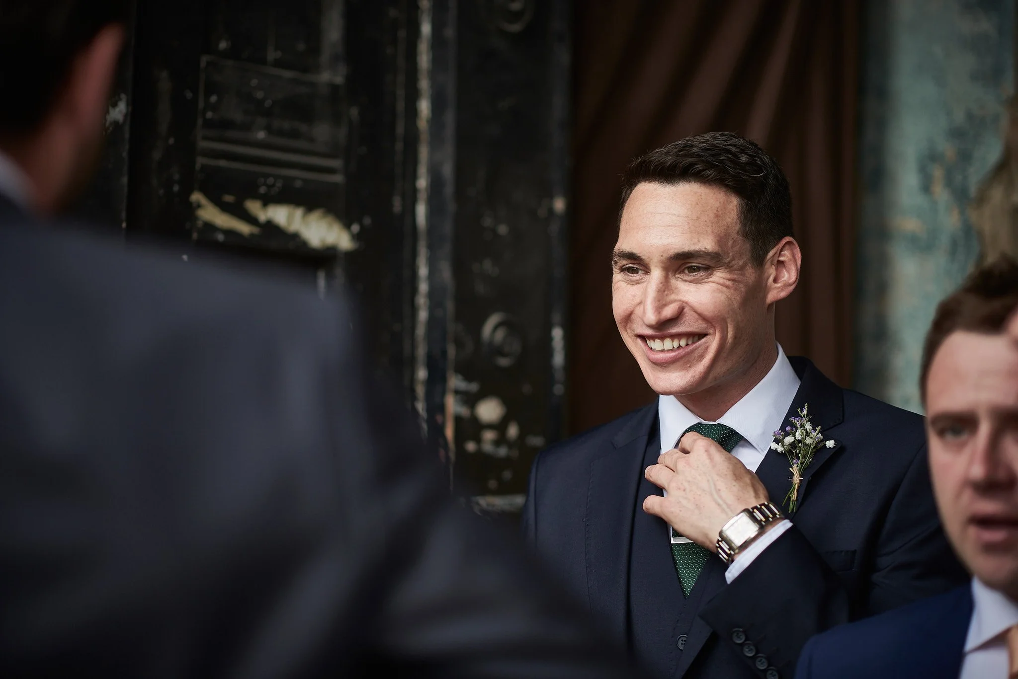A smiling man in a dark suit and white shirt adjusting his green tie at a formal event, with a boutonniere and a watch on his wrist, engaging in conversation with others.