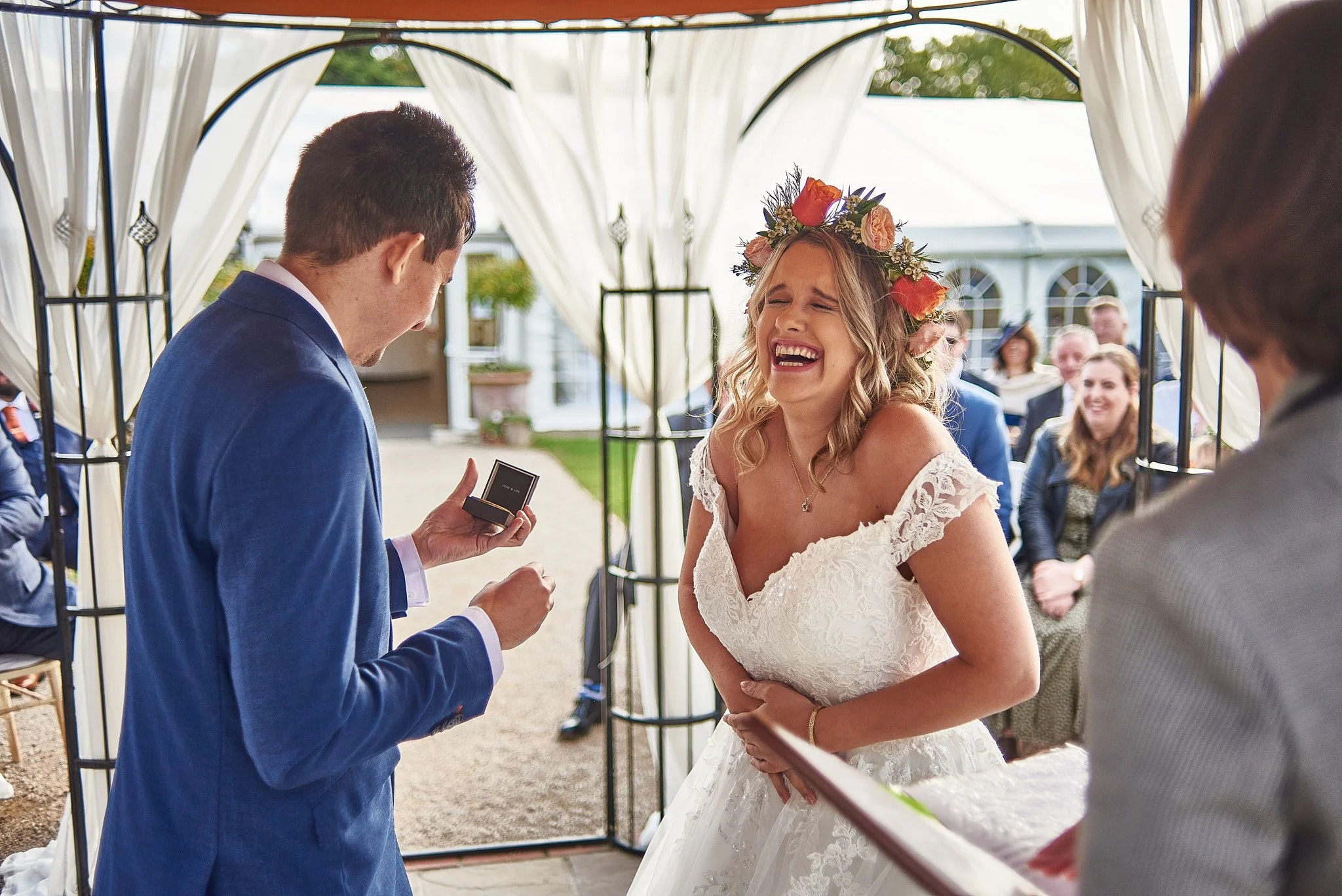 Bride with a floral crown laughing during her wedding ceremony, with a groom in a blue suit holding a ring box in front of her, as guests seated in the background
