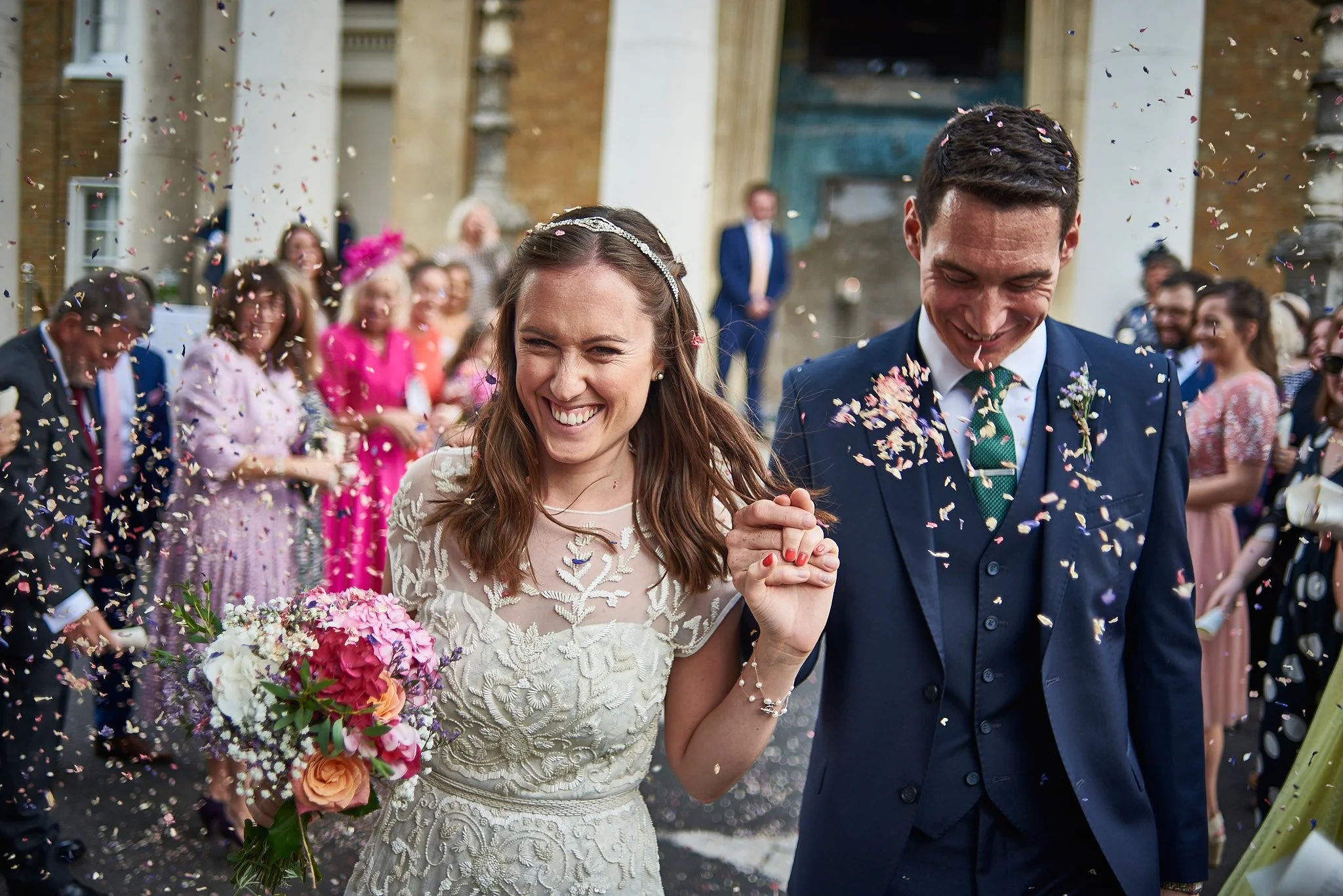 Smiling bride and groom holding hands surrounded by wedding guests and colorful confetti outside a historic building.