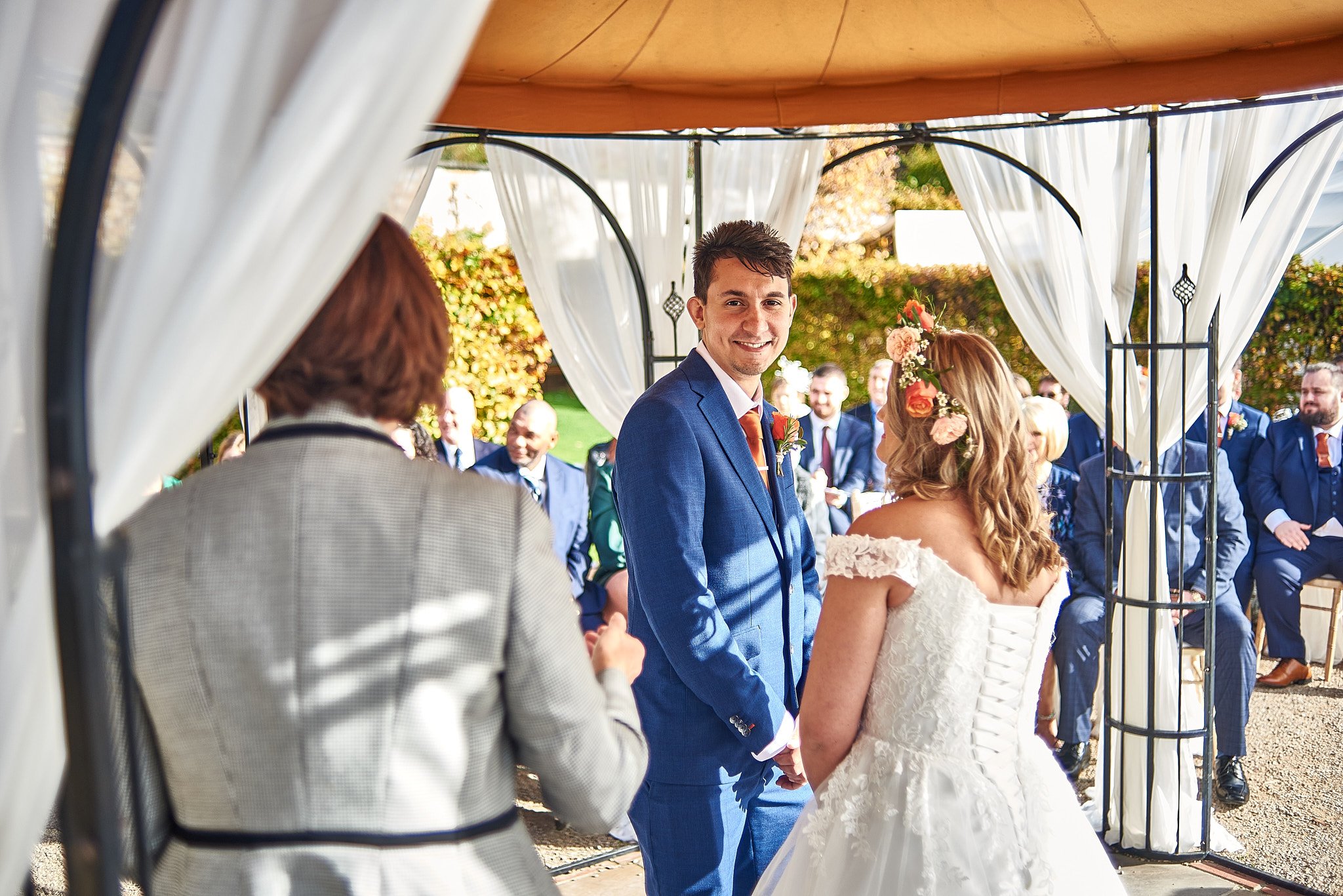 A young groom in a blue suit smiling at his bride during a wedding ceremony outdoors with guests in the background.