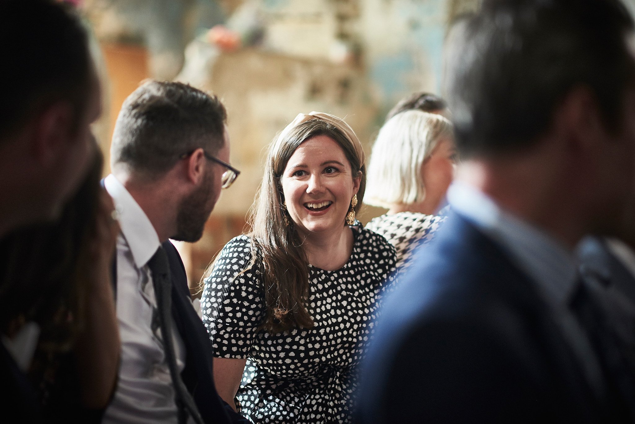 A group of people sitting together at a social gathering, with a smiling woman wearing a black and white polka dot dress in focus.