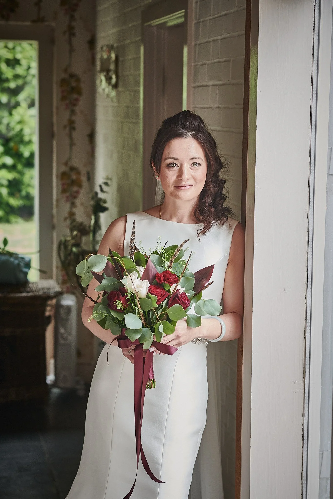 Woman in a white dress holding a bouquet of red, white, and green flowers, standing in a doorway.
