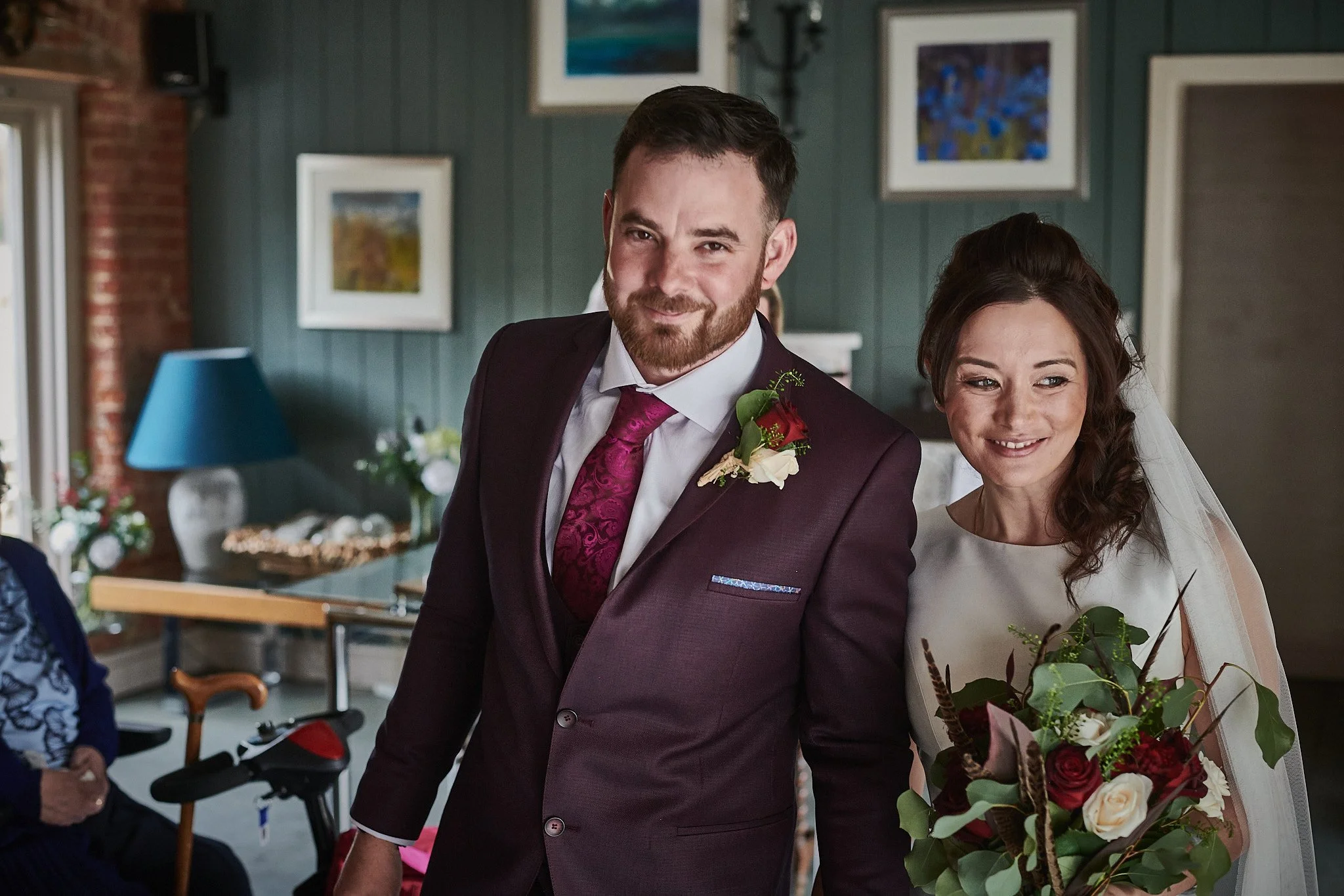 A bride and groom smiling inside a decorated room during their wedding day. The groom is wearing a dark suit with a purple tie and a boutonniere, while the bride is holding a bouquet of flowers and wearing a white wedding dress with a veil.