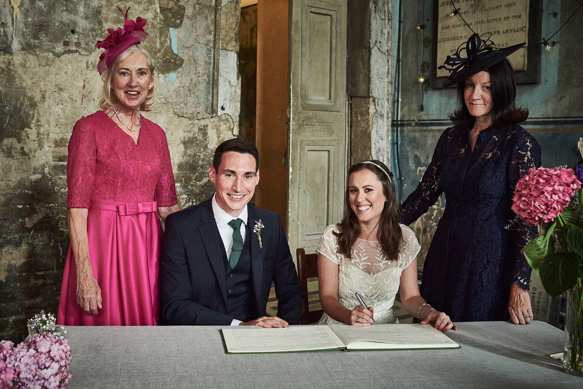 A bride and groom seated at a table signing a marriage register, surrounded by two women wearing fancy hats, in a rustic setting with aged walls and floral arrangements.