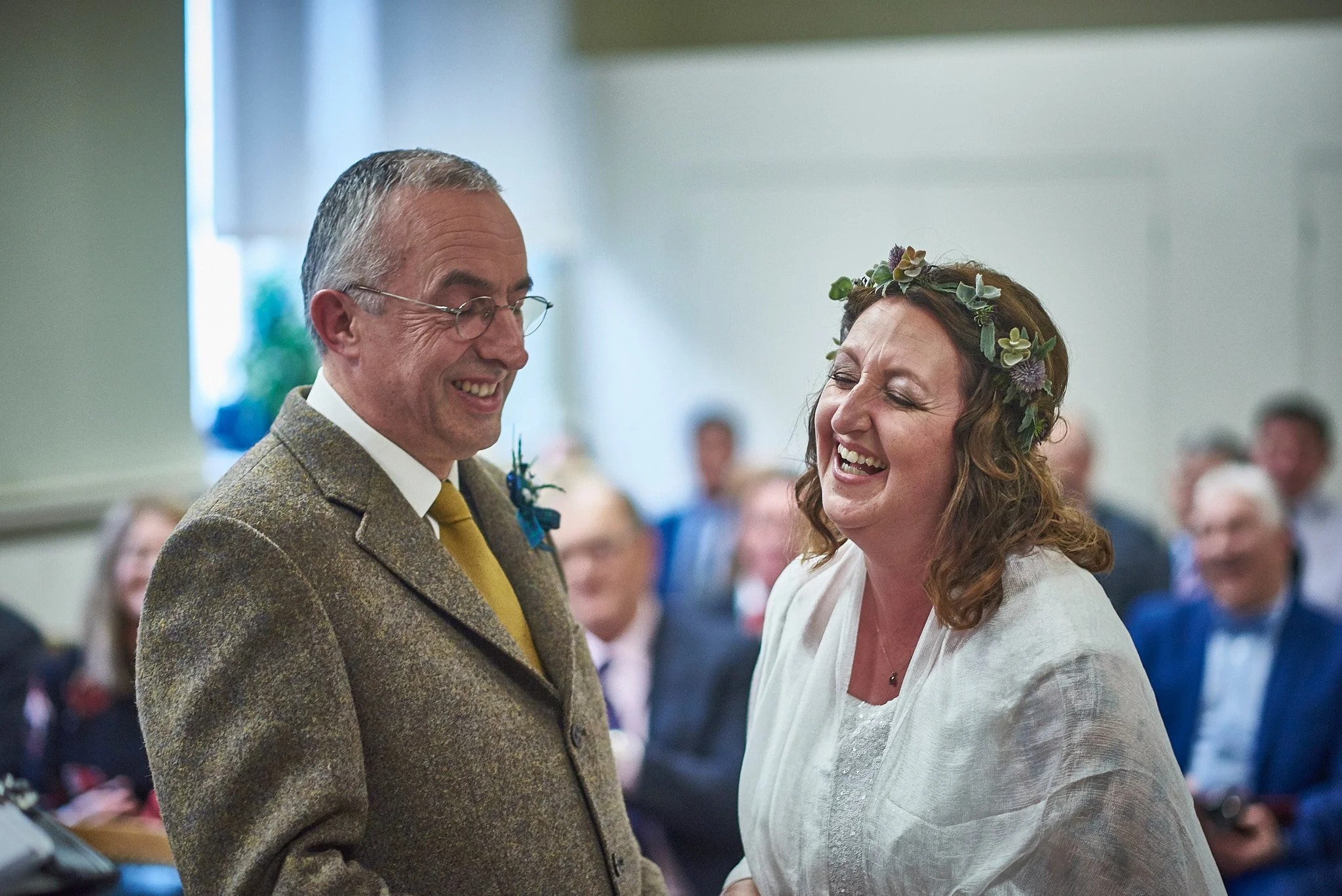 A man and woman smiling at each other during a wedding ceremony. The woman is wearing a floral crown and a white dress, while the man is wearing glasses, a brown tweed suit, and a yellow tie.