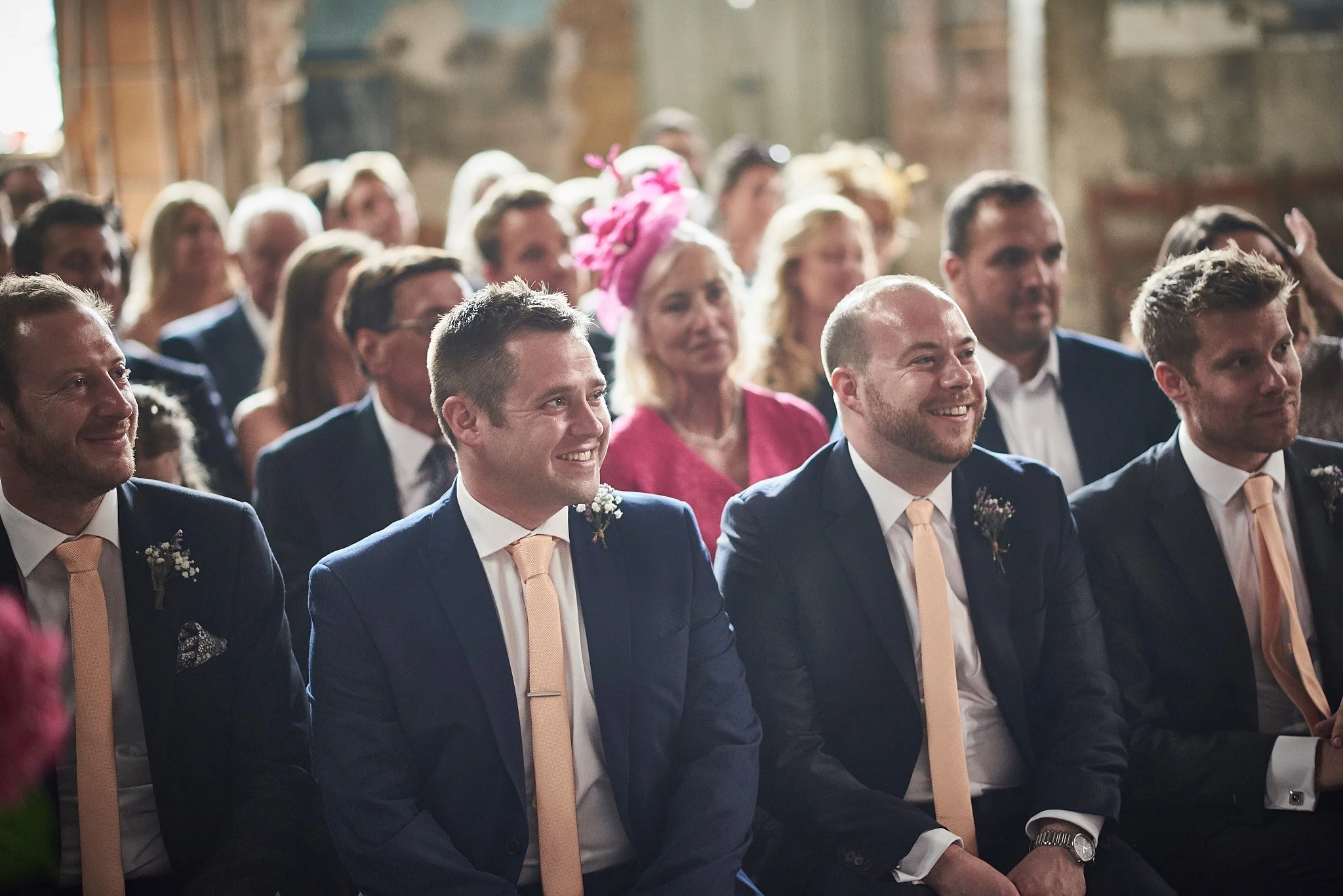 A group of men in suits with peach ties sitting and smiling at a wedding ceremony.