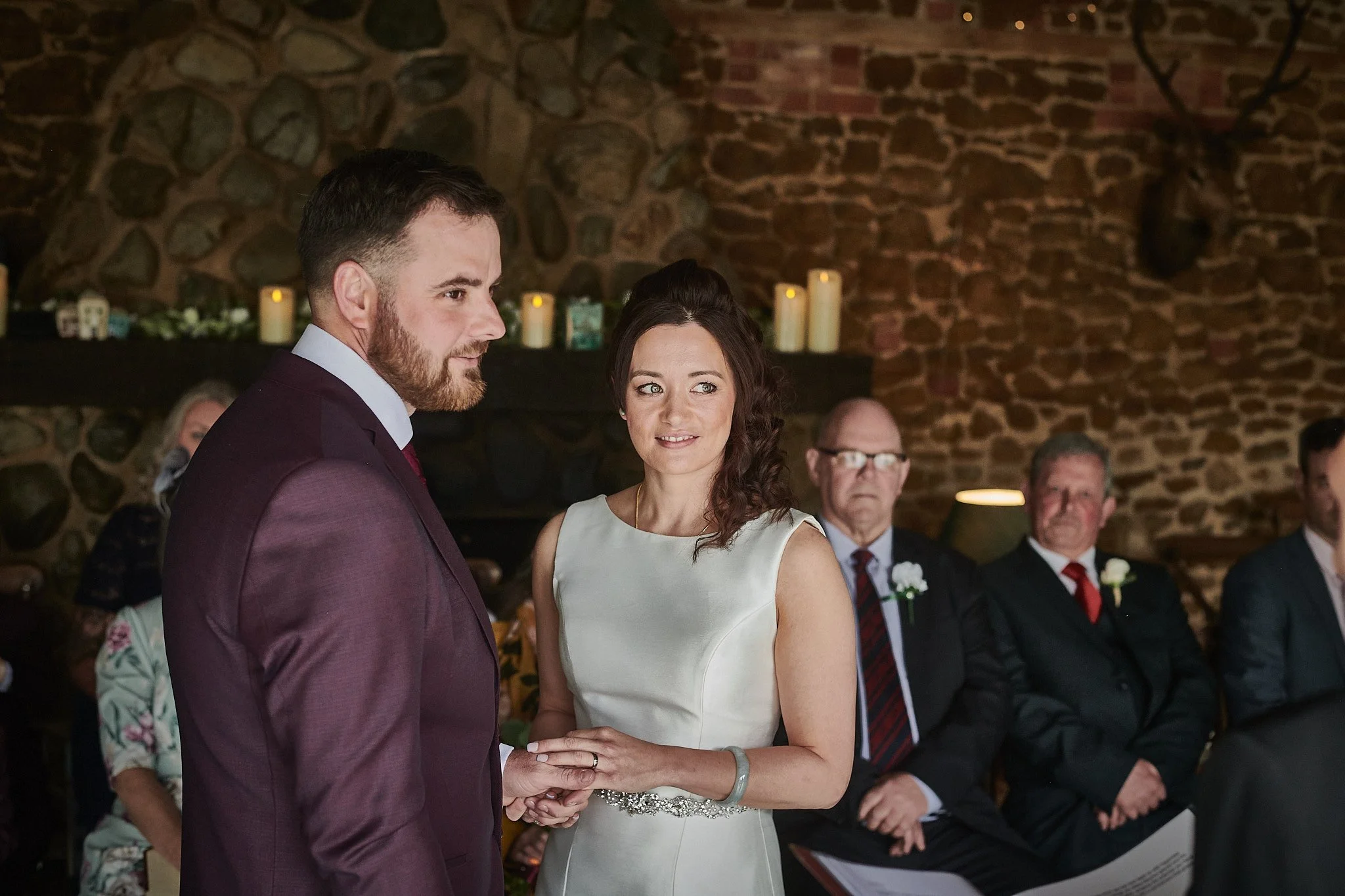 A bride and groom exchanging vows during a wedding ceremony in front of seated guests, inside a rustic brick-walled venue with candles on the mantle.