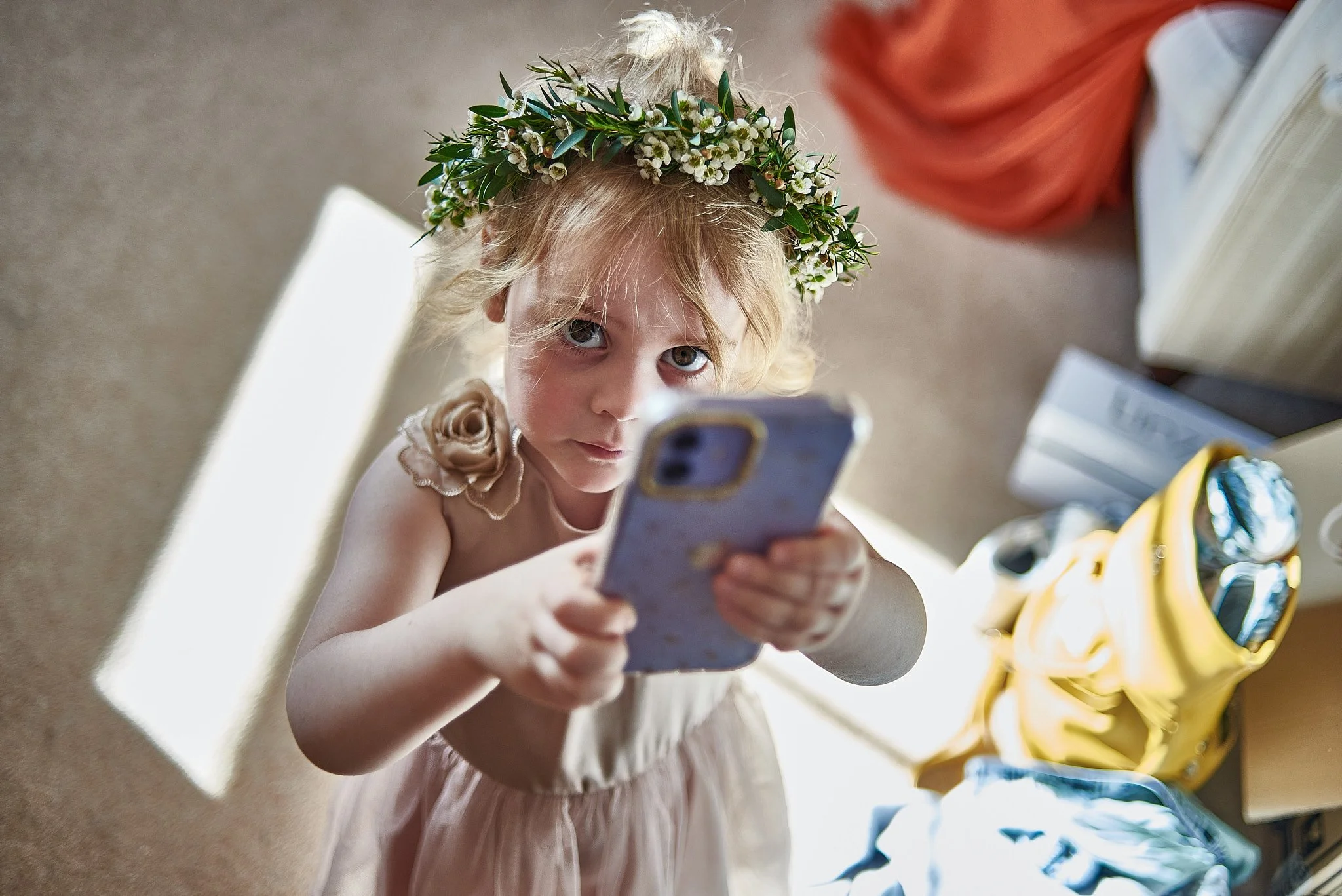 A young girl with blonde hair wearing a flower crown and a dress, looking up at the camera while holding a smartphone, surrounded by bags and boxes.