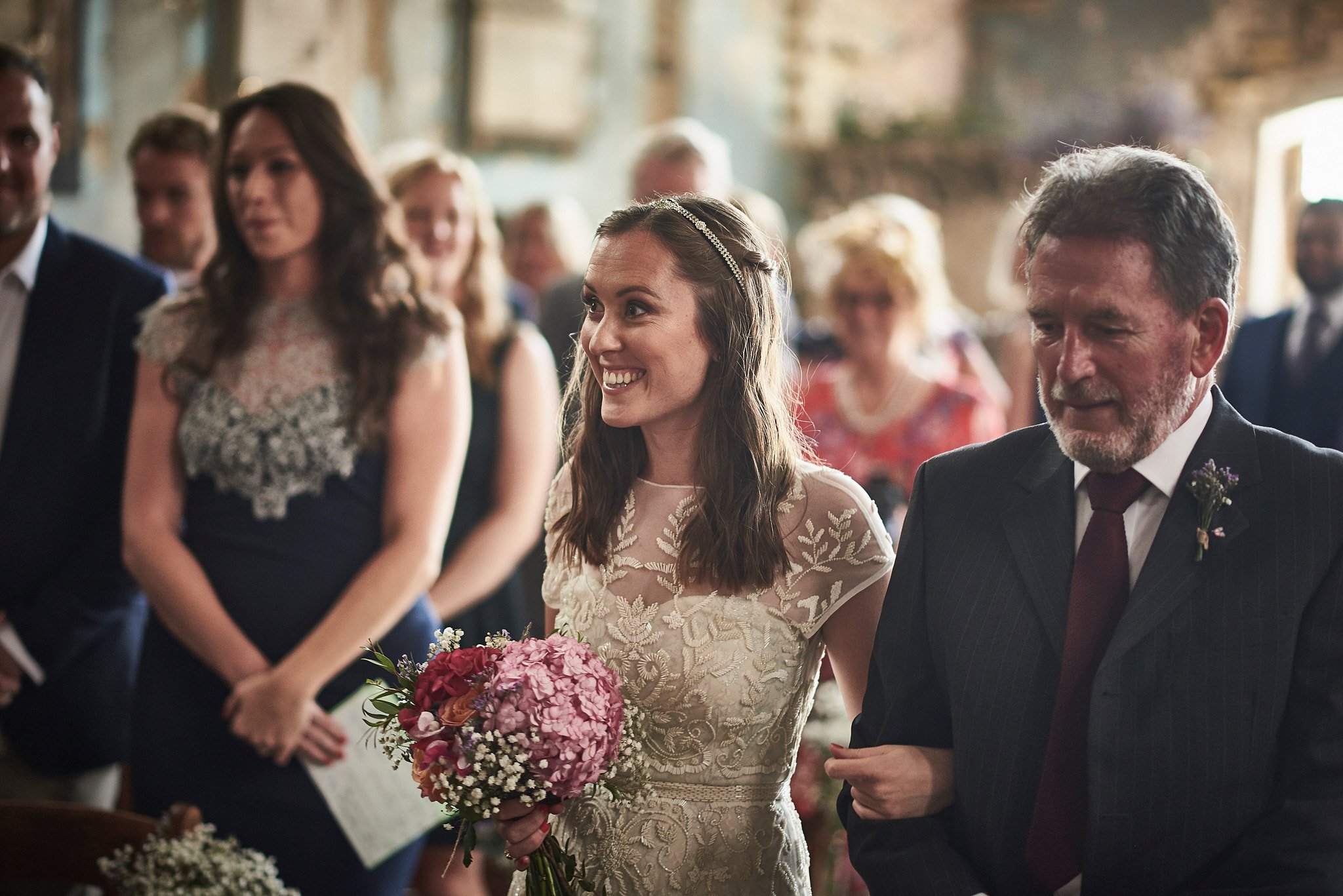 Bride smiling and holding a bouquet, walking down the aisle with an older man, possibly her father, during a wedding ceremony surrounded by guests.