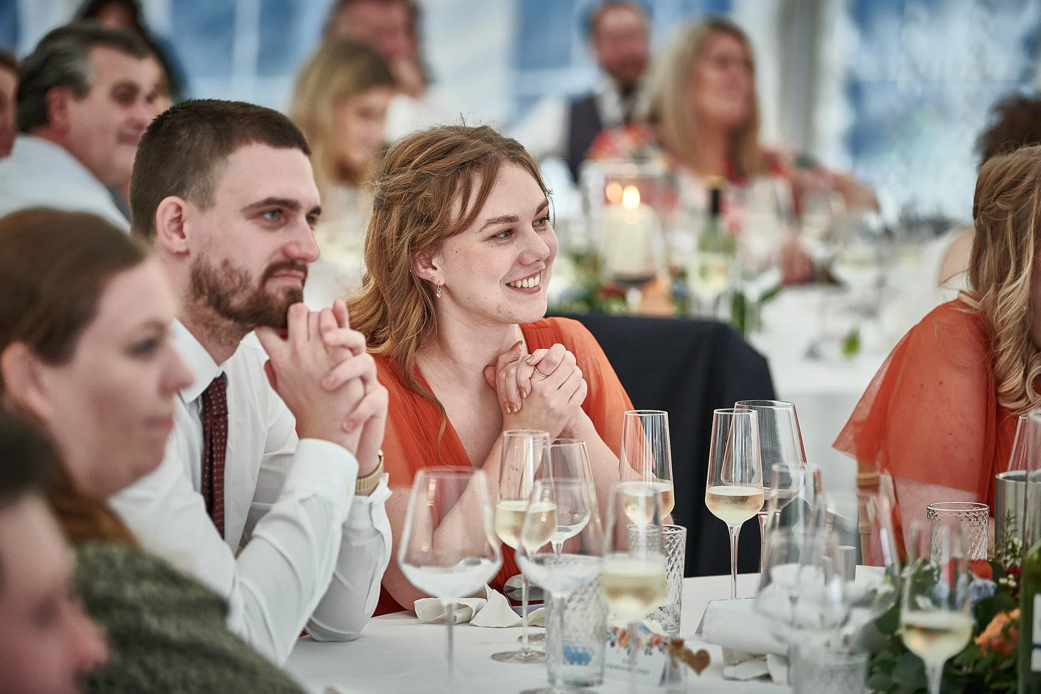 People sitting at a table, smiling and clapping, with glasses of champagne and floral decorations at a formal event or celebration.