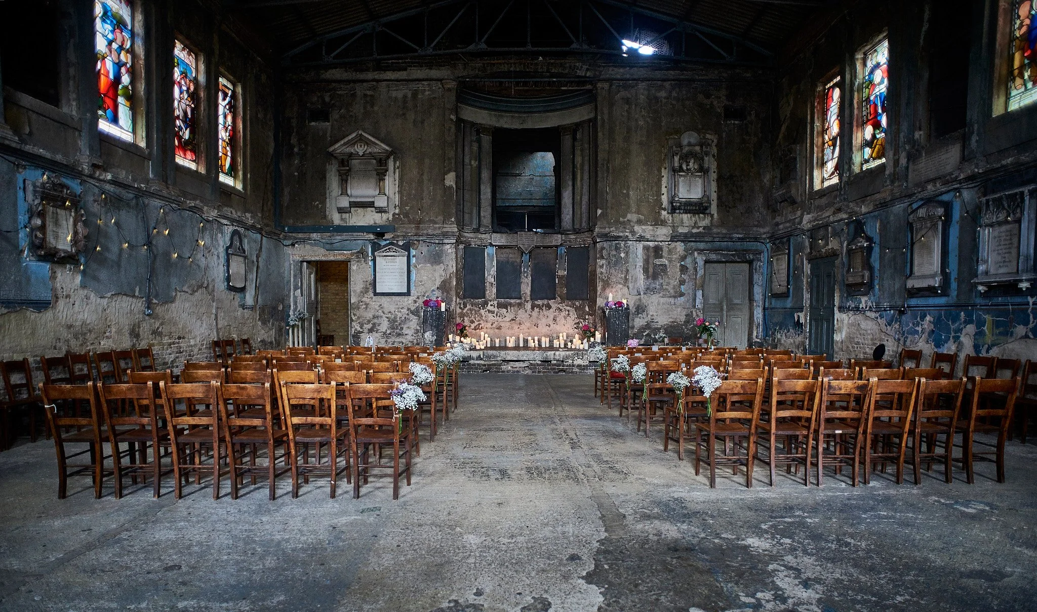 Empty indoor wedding or event venue with rustic decor, wooden chairs arranged in rows, floral arrangements, candles, and stained glass windows.