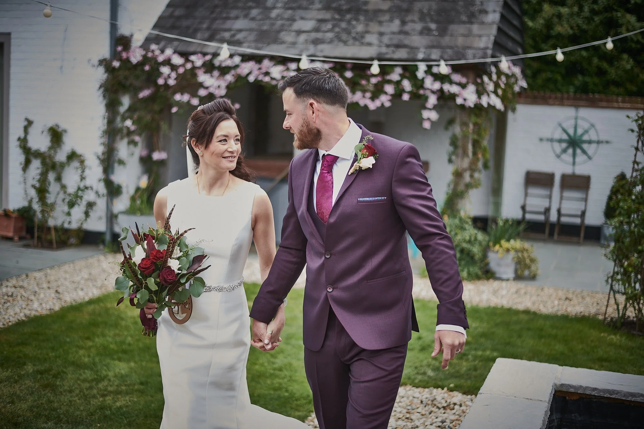 A bride and groom holding hands and smiling at each other in an outdoor garden setting on their wedding day.