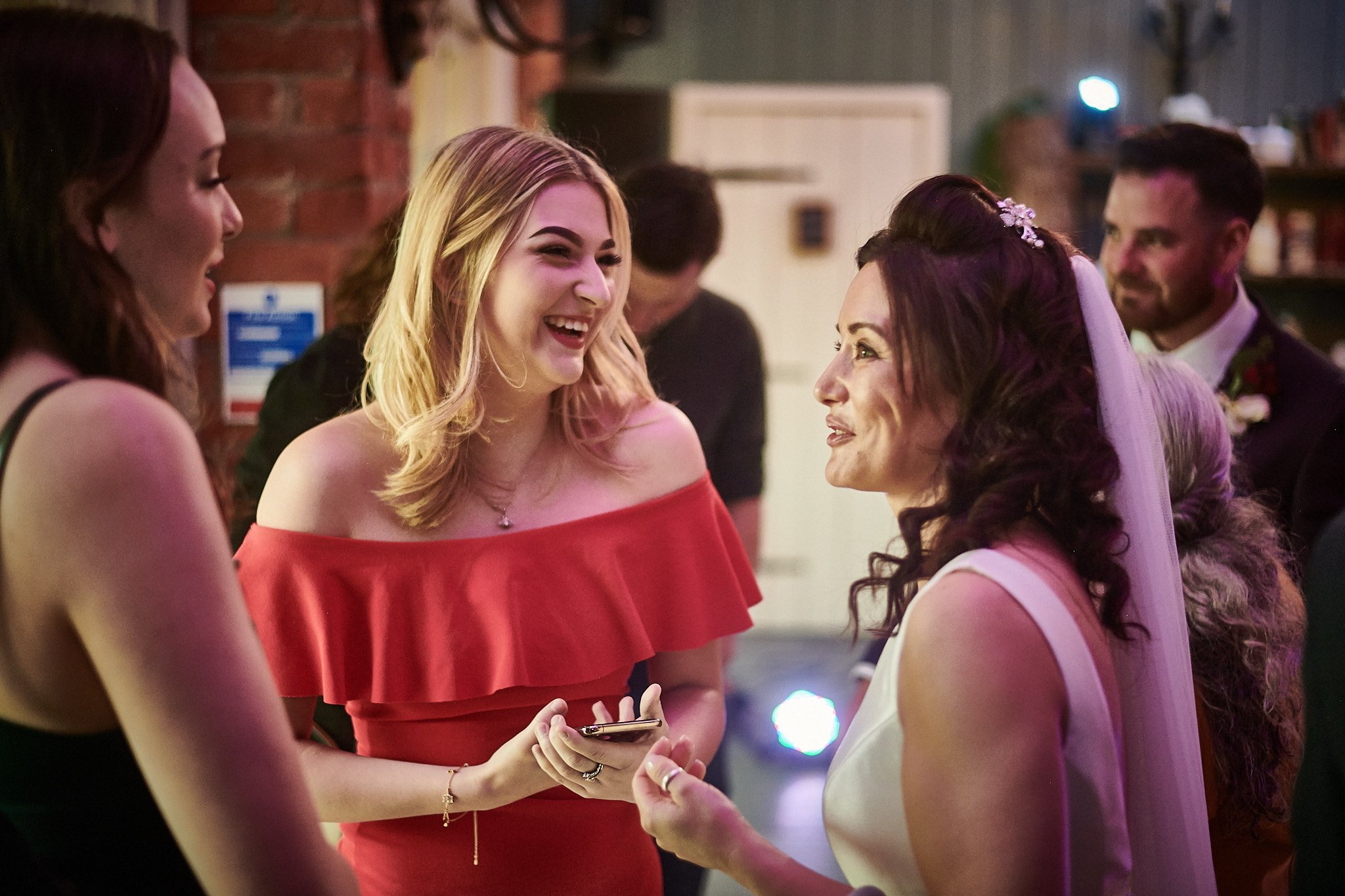 A bride and a woman in a red off-shoulder dress are smiling and talking at a wedding reception, with other guests in the background.