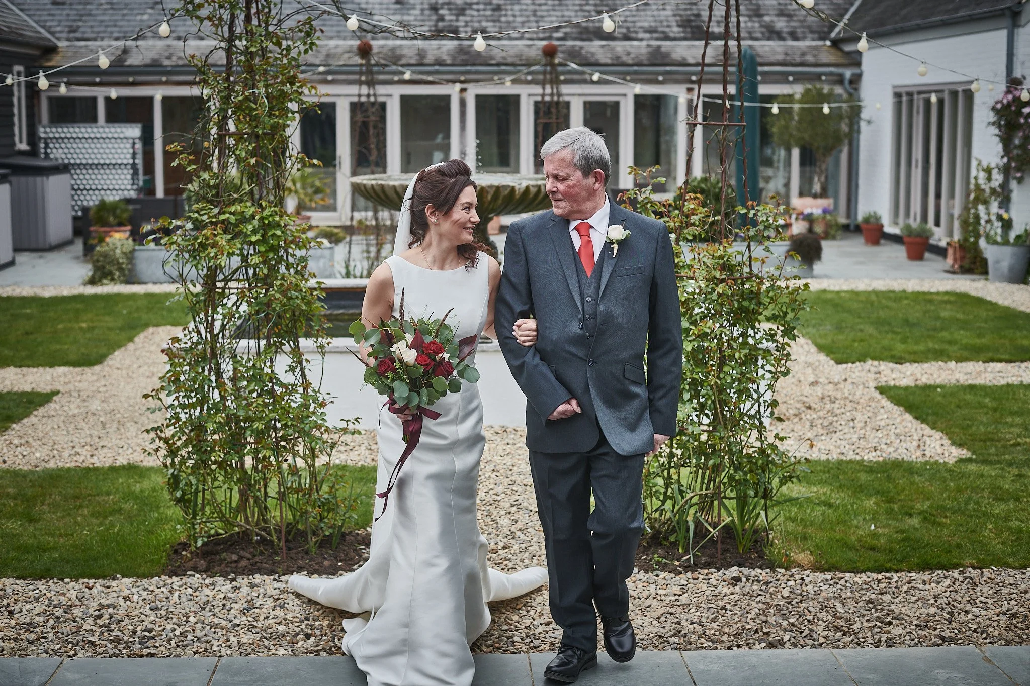 A bride in a white wedding dress holding a bouquet of red and white flowers, walking arm-in-arm with an older man in a suit, outside in a garden setting decorated with string lights and greenery.