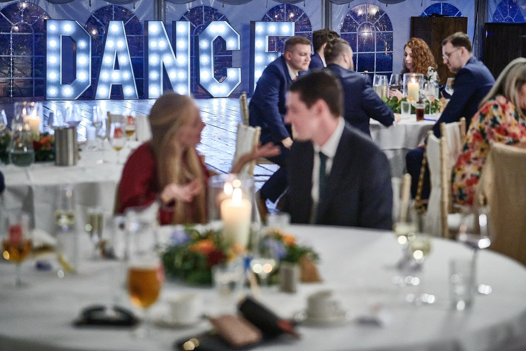 People sitting at decorated tables in a ballroom with a large illuminated 'DANCE' sign in the background.