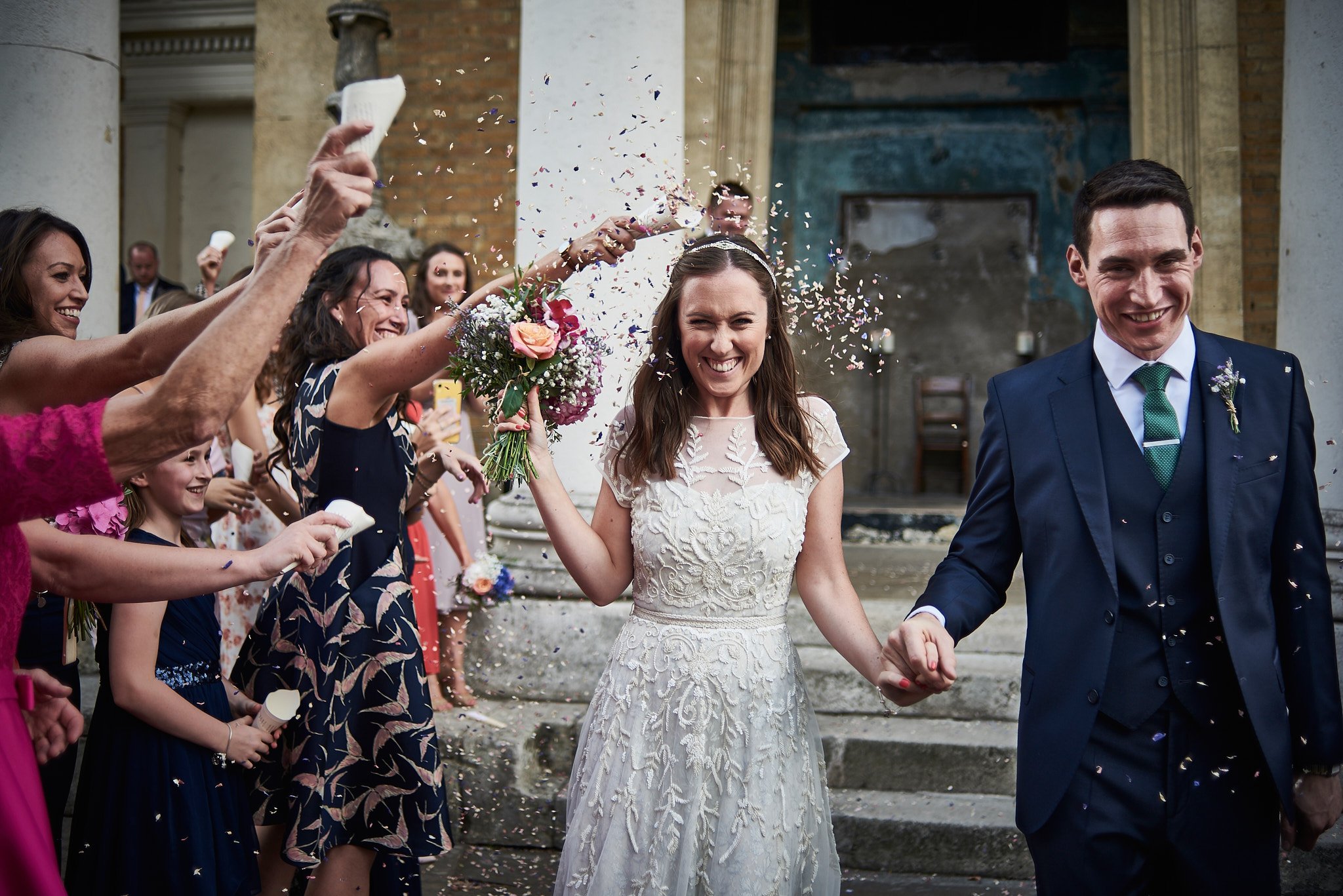 Bride and groom smiling and walking hand in hand as guests shower them with confetti outside a building with columns.