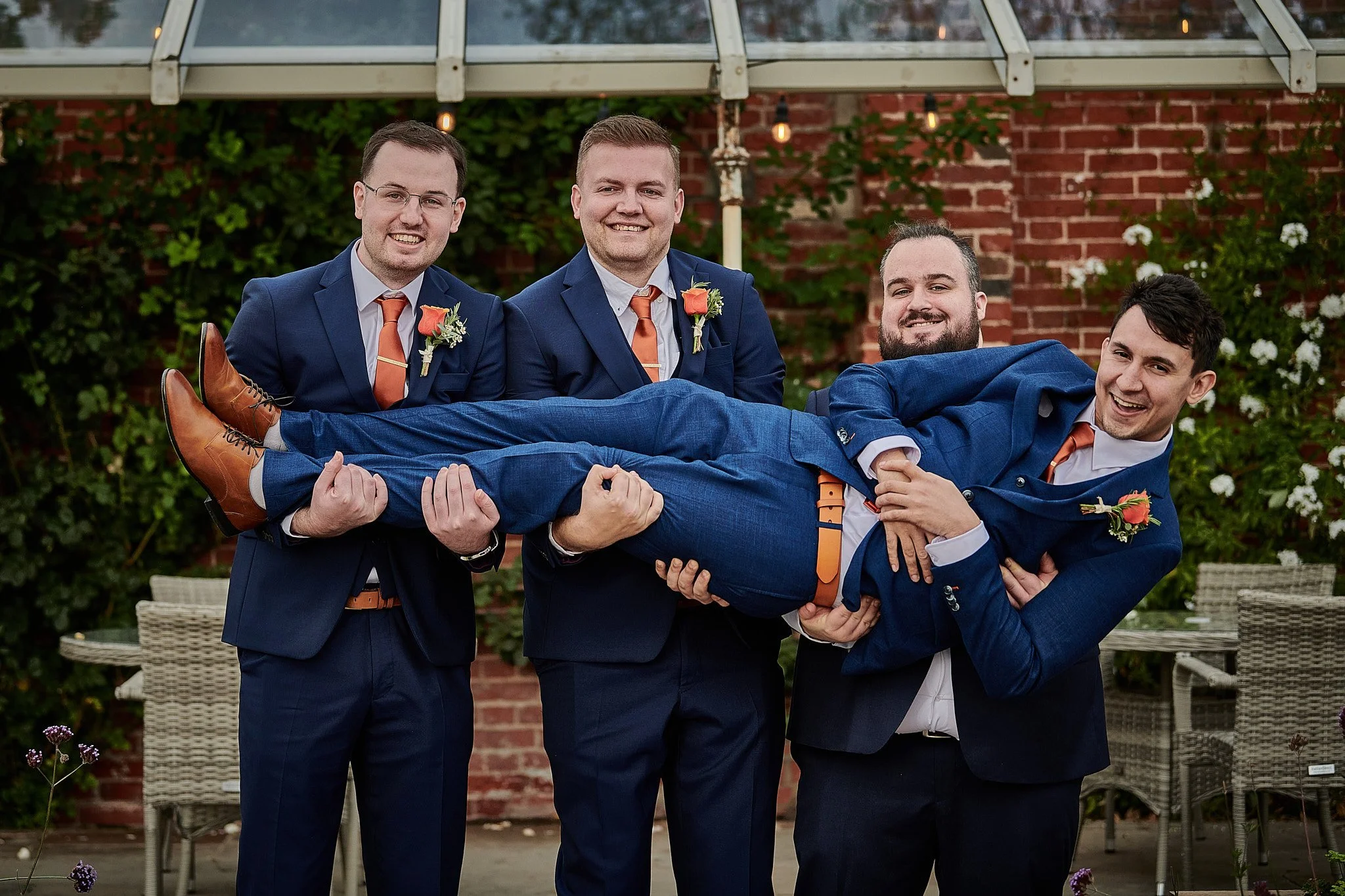 Four men in suits, three holding a fourth man horizontally. All are smiling; the three standing are in navy suits with orange ties and corsages. The man being held is in a matching navy suit and brown shoes, smiling with his arms crossed. The backgro