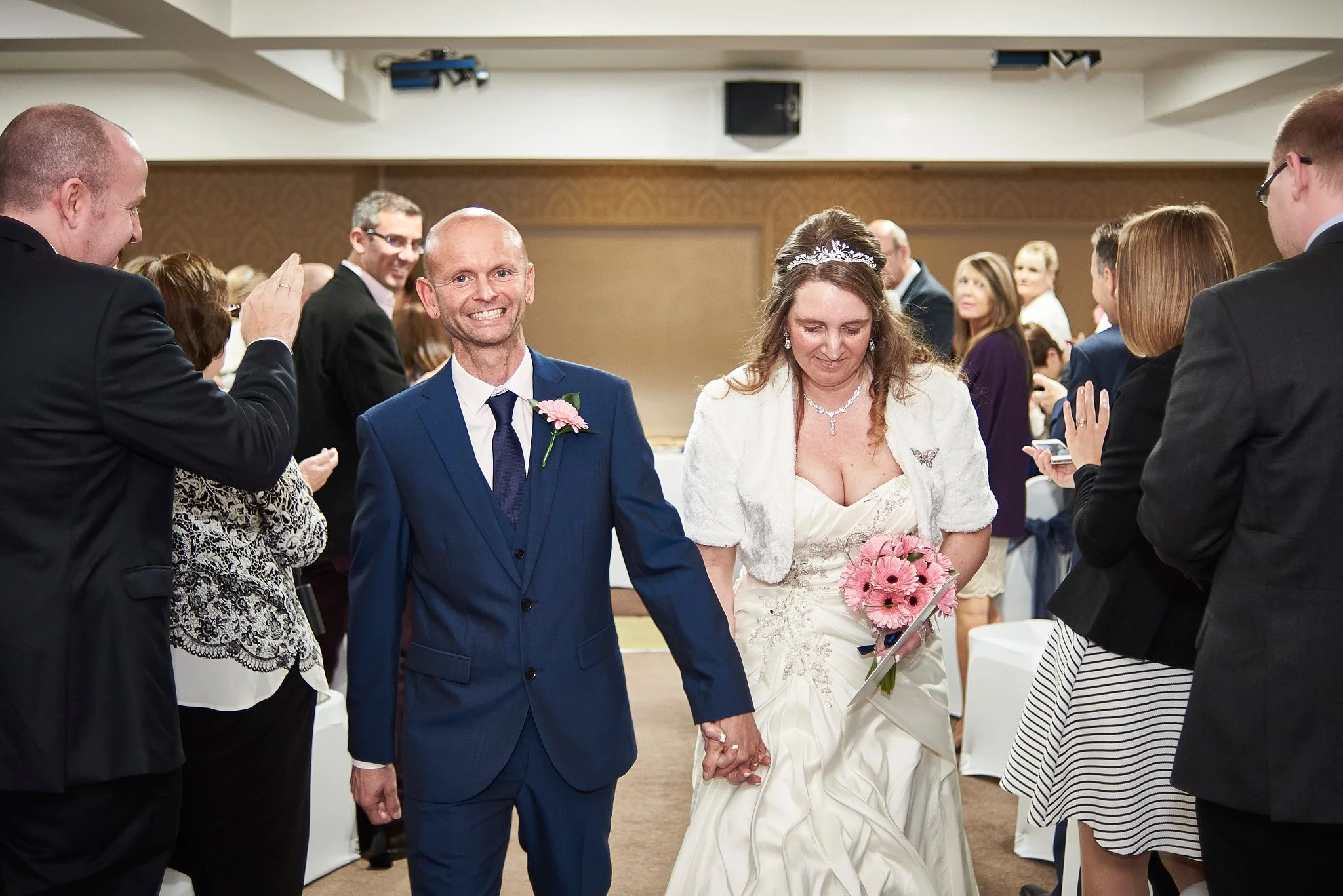 A wedding reception with a bride and groom walking hand in hand, surrounded by guests applauding and taking photos in a banquet hall.