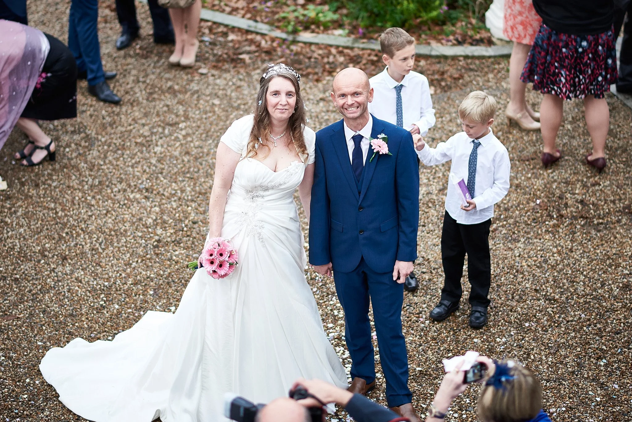 Bride in a white wedding gown holding a pink bouquet standing next to a man in a blue suit with a pink ribbon boutonniere, during a wedding ceremony, with children and guests in the background.