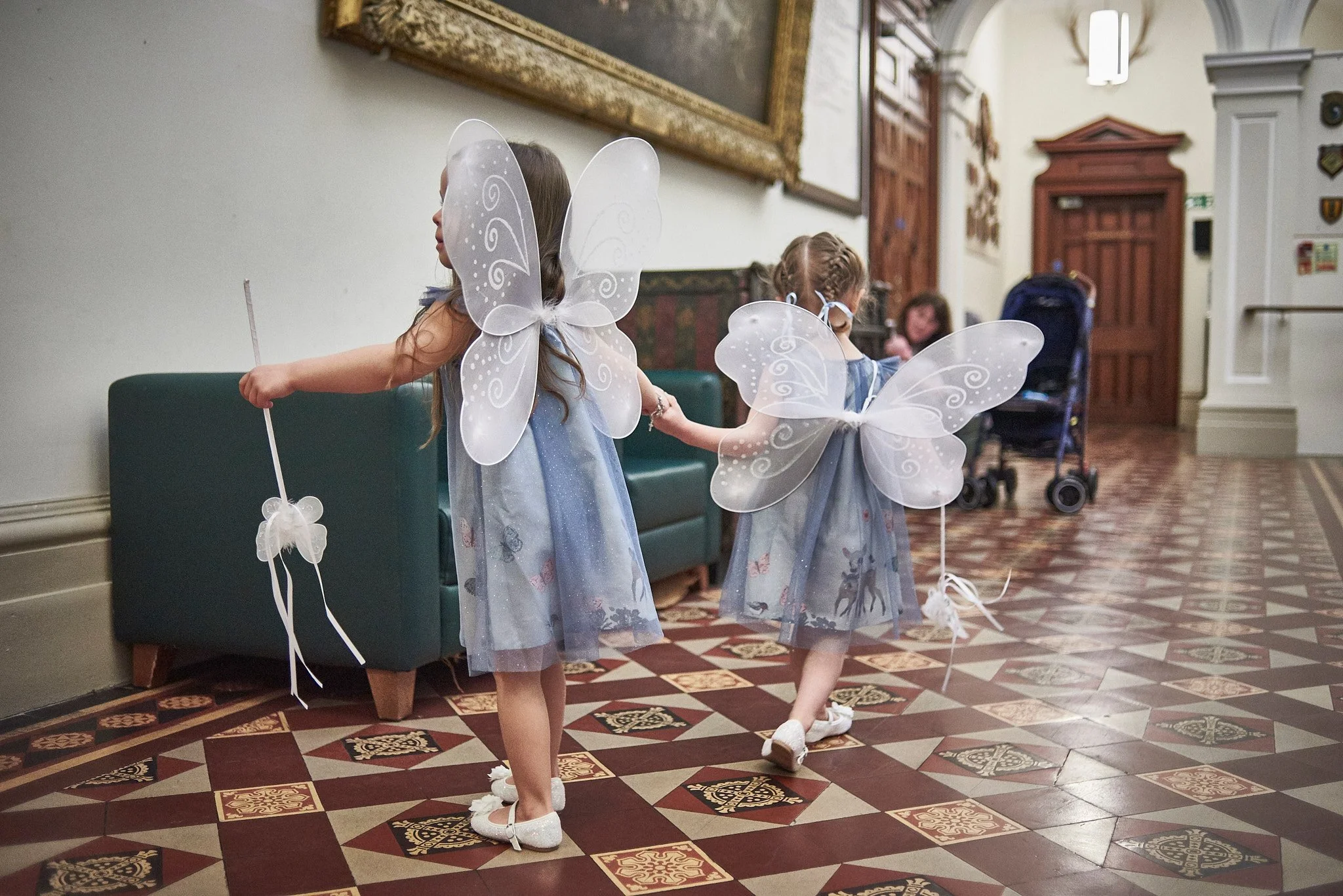 Three young girls dressed as fairies with fairy wings and wands, holding hands, walking through an indoor hallway with a patterned tile floor.