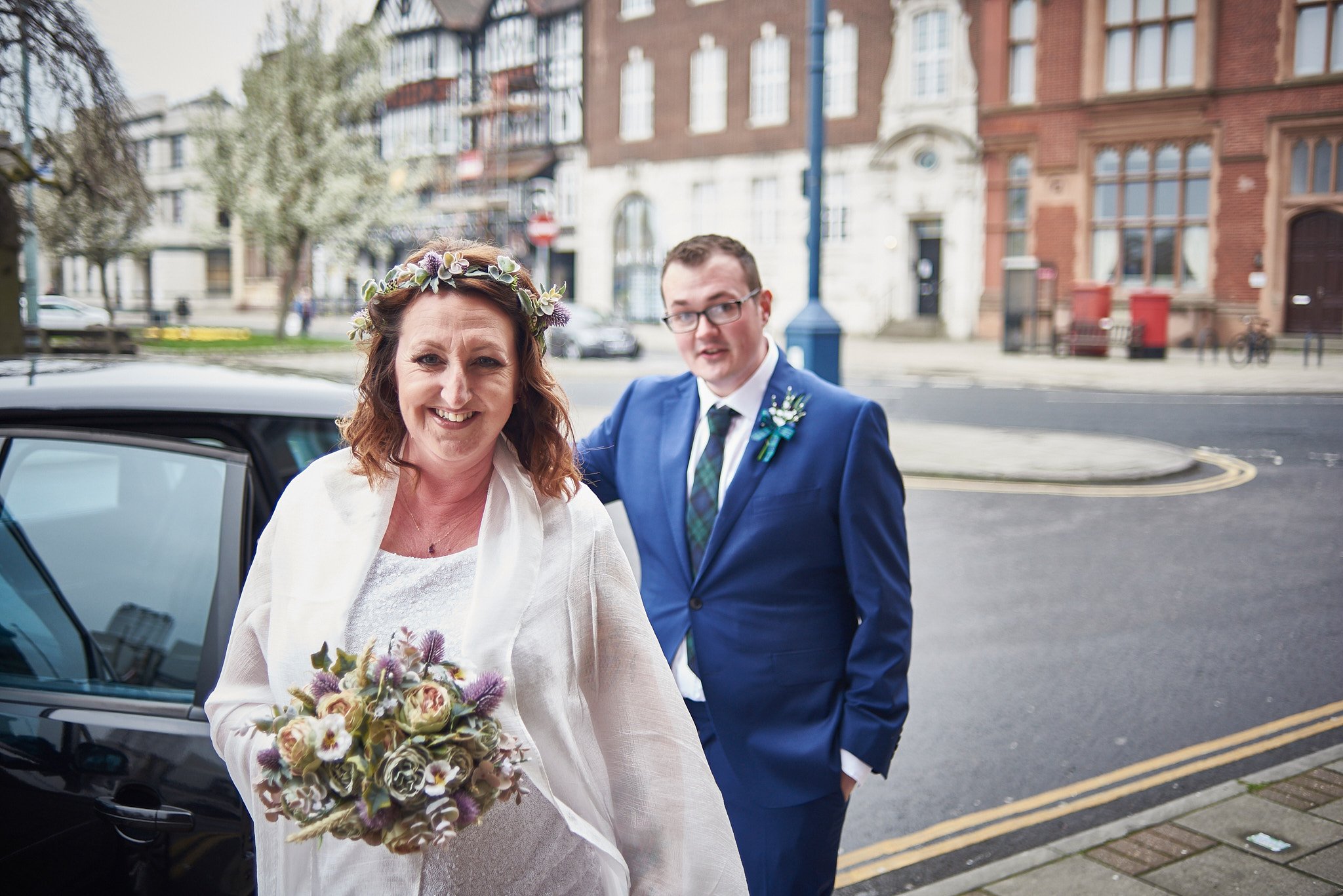 A woman in a wedding dress holding a bouquet and wearing a flower crown, smiling, with a man in a blue suit and tie standing behind her outside on a city street.