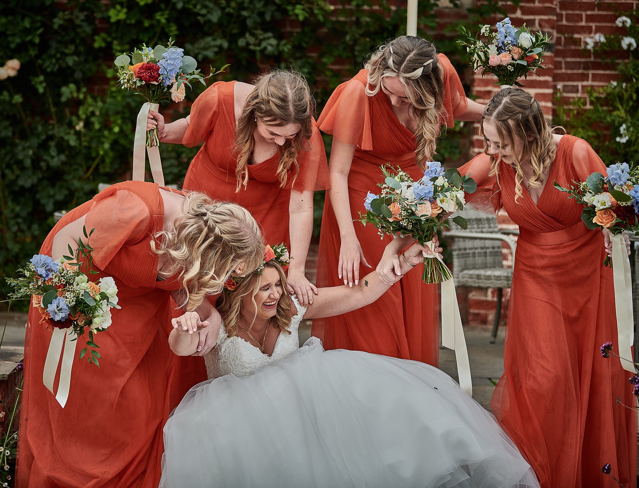 Bride in white lying on the ground with five bridesmaids in orange dresses surrounding her in a garden, all holding bouquets and laughing.