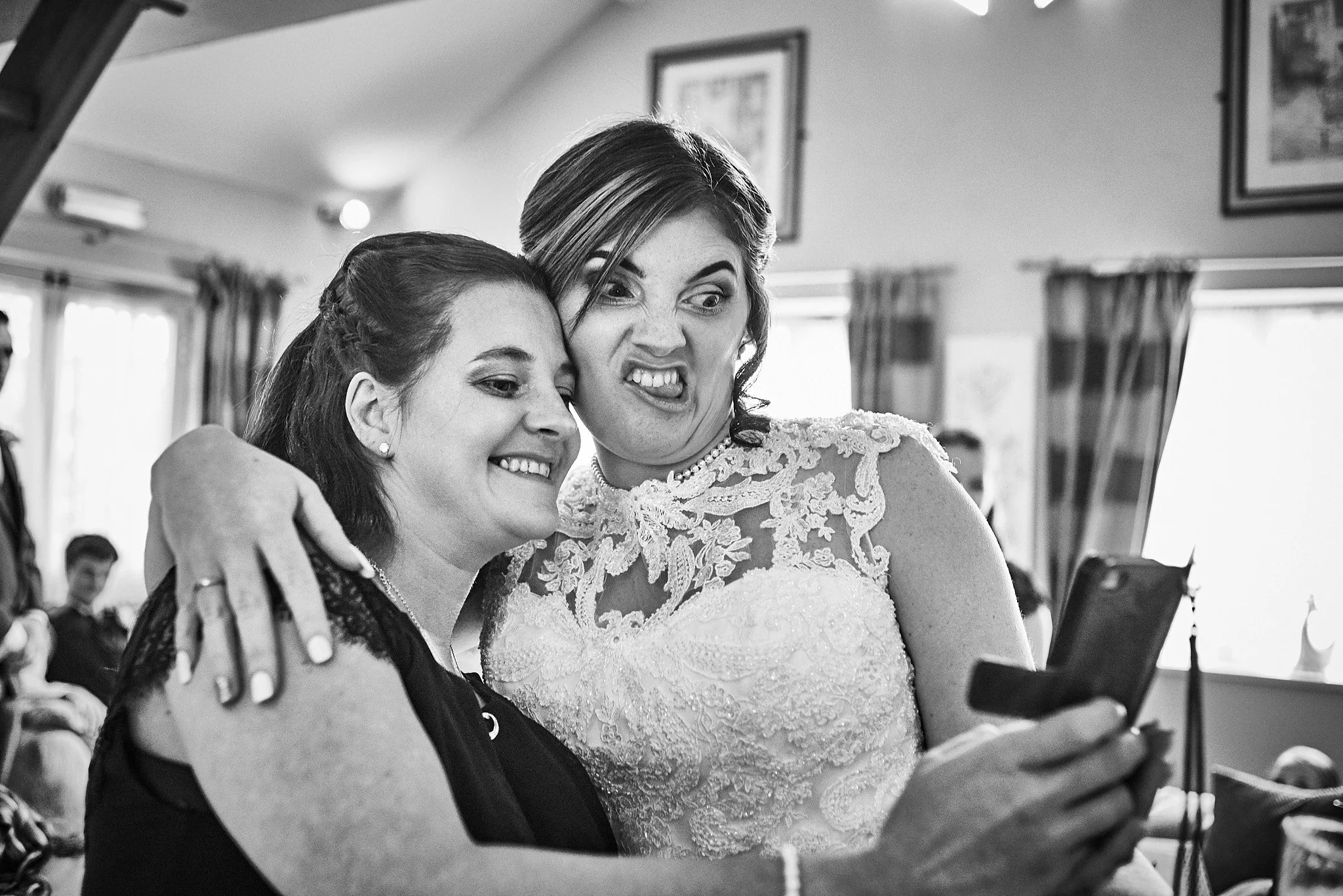 Black and white photo of two women, one in a wedding dress, looking at a phone together and making funny faces. The woman in the wedding dress has her arm around the other woman, both smiling and posing for the camera. In the background, there are ot