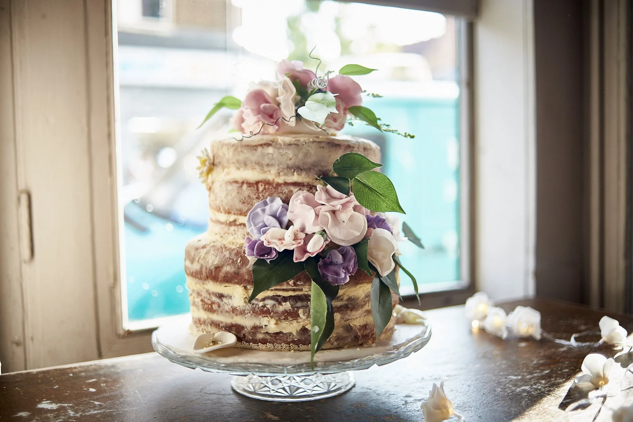 A rustic, semi-naked wedding cake decorated with pink, white, and purple flowers and green leaves, sitting on a glass cake stand near a window.
