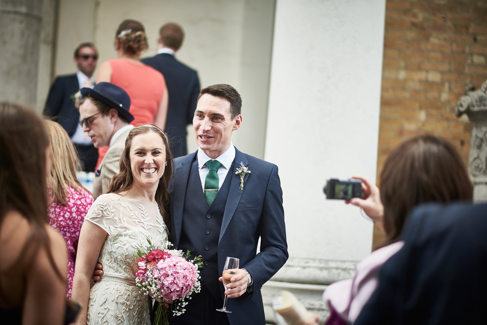 A bride and groom smiling at a wedding reception, surrounded by guests, with one person taking a photo.
