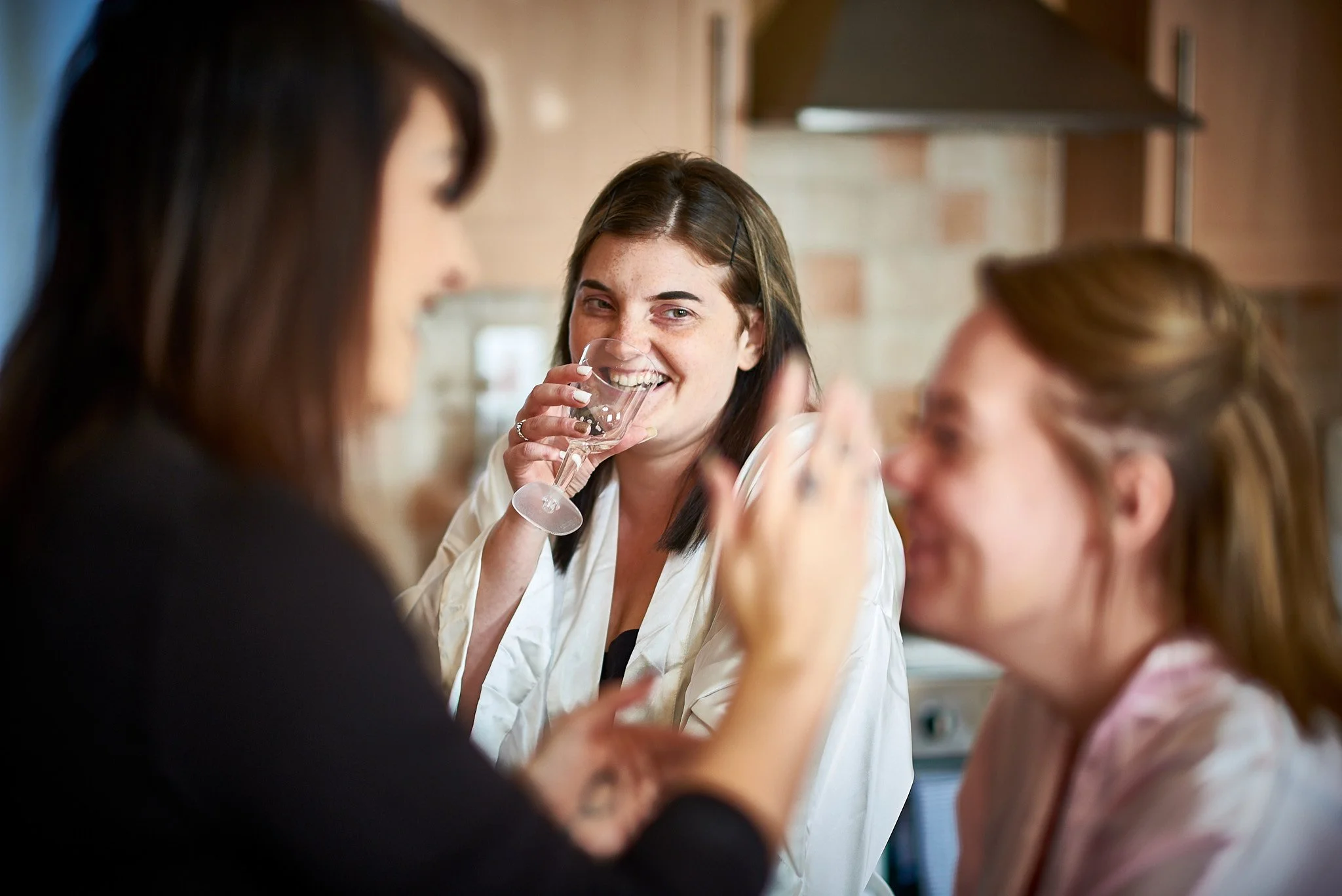 Three women having a conversation, one drinking from a wine glass, in a cozy indoor setting with brick walls.