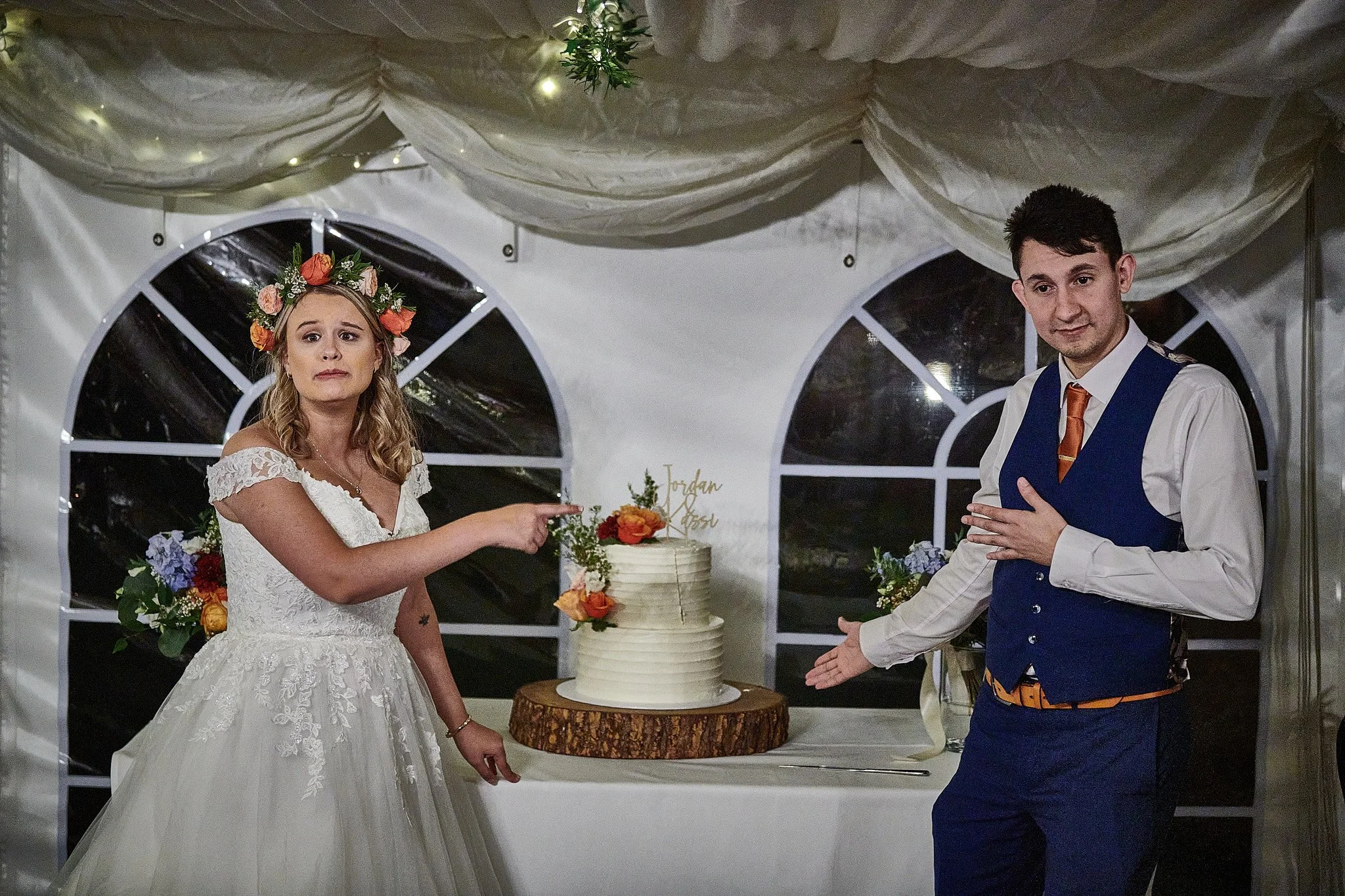 A bride and groom at a wedding reception, the bride wearing a floral crown and a white lace gown, pointing to a wedding cake, the groom standing nearby with one hand on his chest, inside a decorated tent.