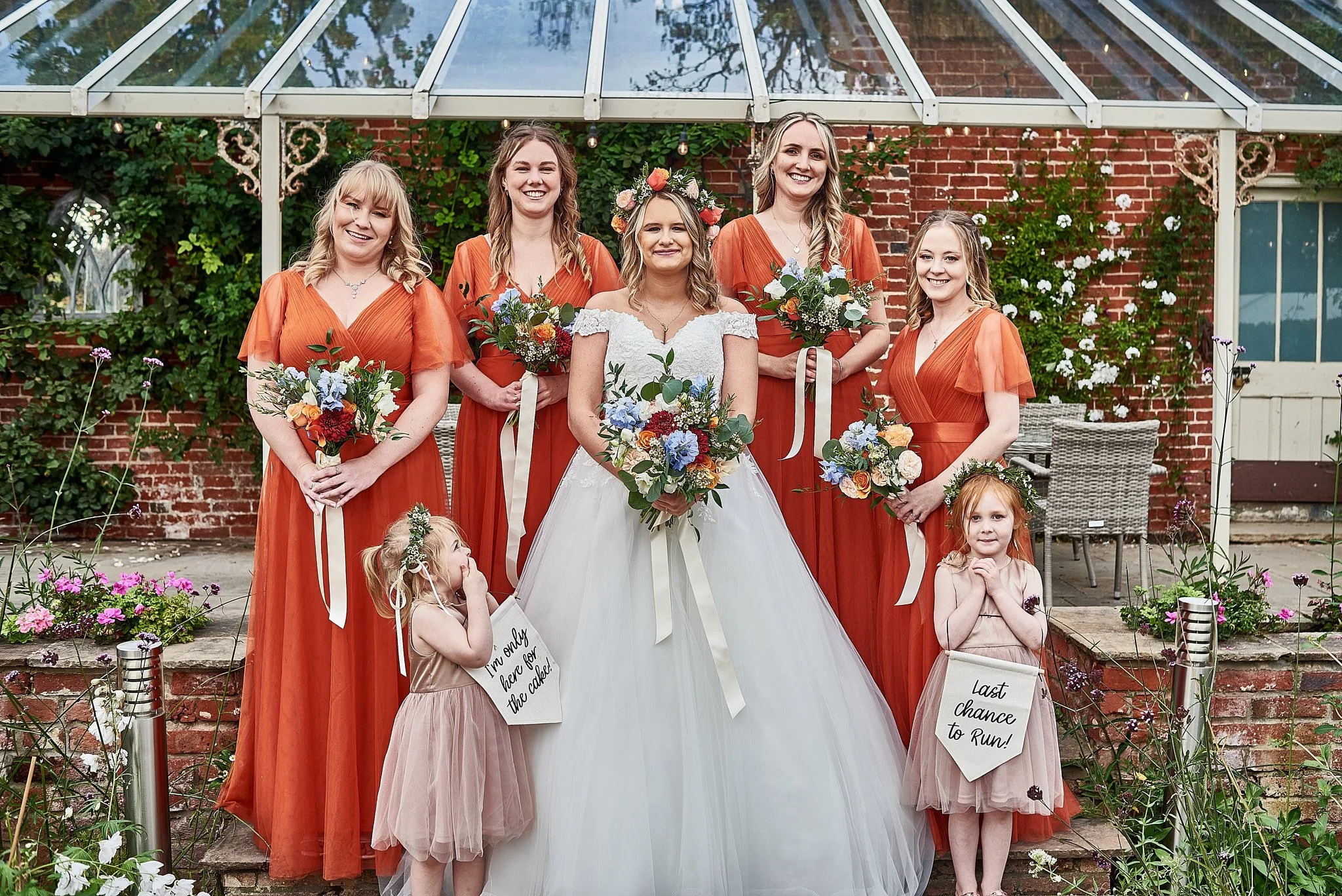 A bride in a white wedding gown with a floral crown stands with six women in orange bridesmaid dresses and two young flower girls outdoors in a garden setting.
