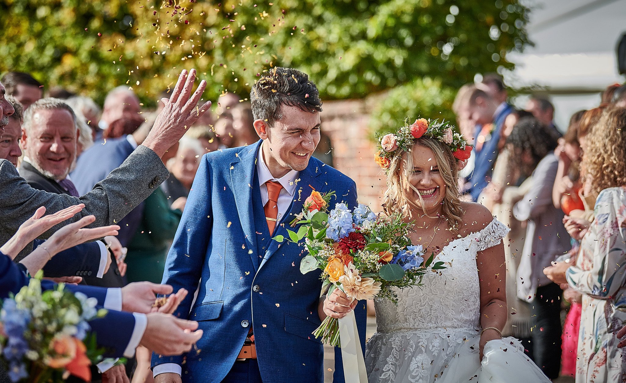 Happy newlywed couple celebrating wedding with guests, confetti, and flowers outdoors.