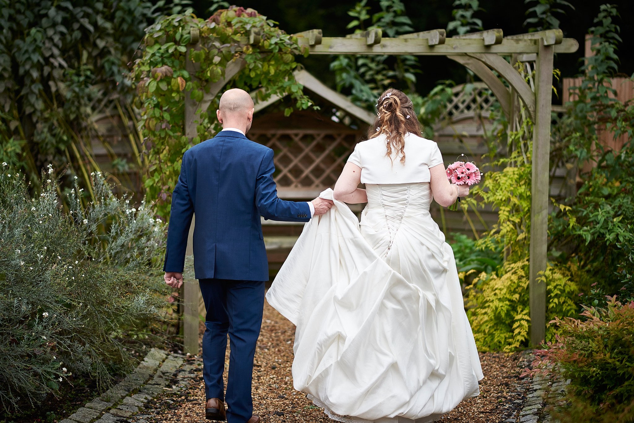 A bride and groom walk arm-in-arm through a garden archway, with the groom holding the train of the bride's white wedding dress. The bride holds a bouquet of pink flowers.