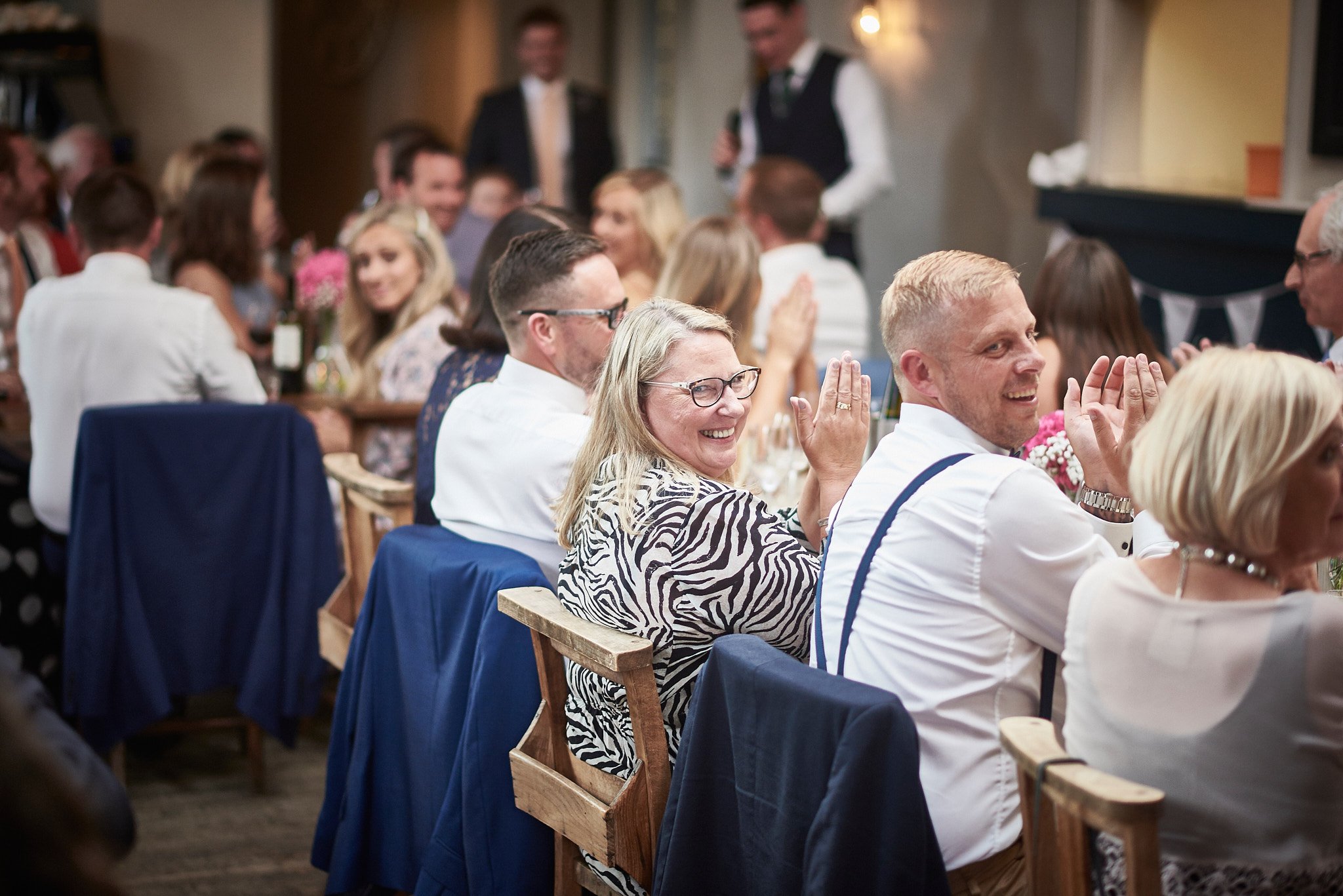 People sitting at tables, smiling, clapping, and enjoying a celebration, with a man speaking at the front, in an indoor setting with warm lighting.