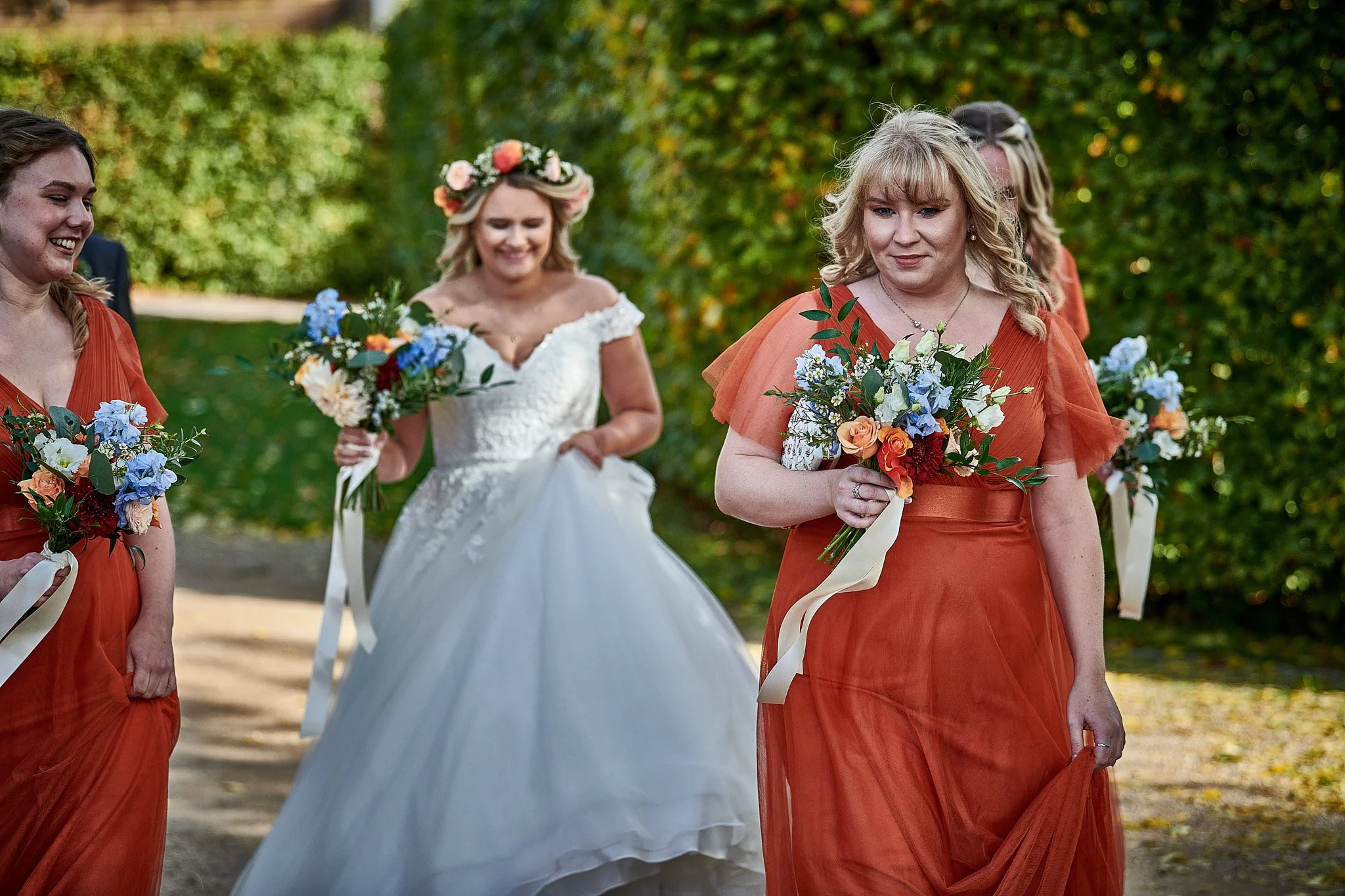 A bride in a white wedding gown walking outdoors with bridesmaids in orange dresses, holding bouquets of colorful flowers, surrounded by greenery.