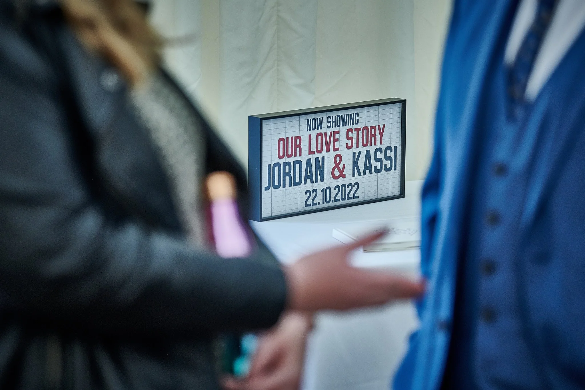 A sign on a table displays information about a movie titled "Our Love Story" starring Jordan and Kassi, showing the date 22.10.2022, with two people in the foreground engaged in conversation.